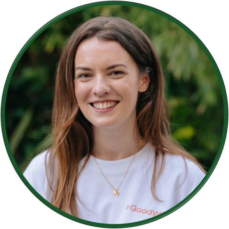 Felicity Halstead  standing outdoors against lush green foliage. She wears a white “GoodWork” T-shirt and gold necklace
