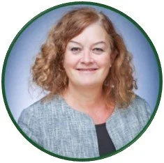 Professional portrait photograph of Sarah with curly auburn hair. She wears a gray patterned blazer over a black top against a blue studio background.