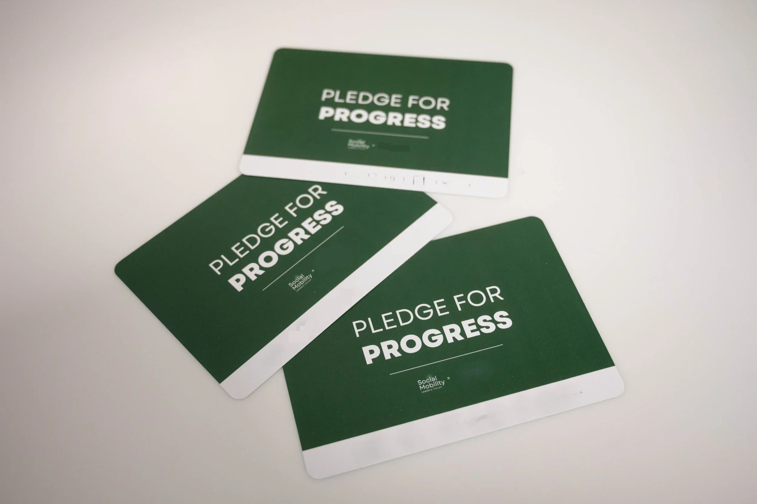 Photograph showing three dark-green pledge cards labeled “PLEDGE FOR PROGRESS,” arranged on a light background.