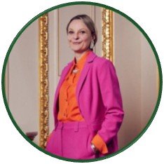 Portrait photograph of a Sarah posing indoors beside ornate gold-trimmed mirrors. She wears a vivid fuchsia suit over an orange blouse, with one hand in a pocket and elegant architectural décor framing her.