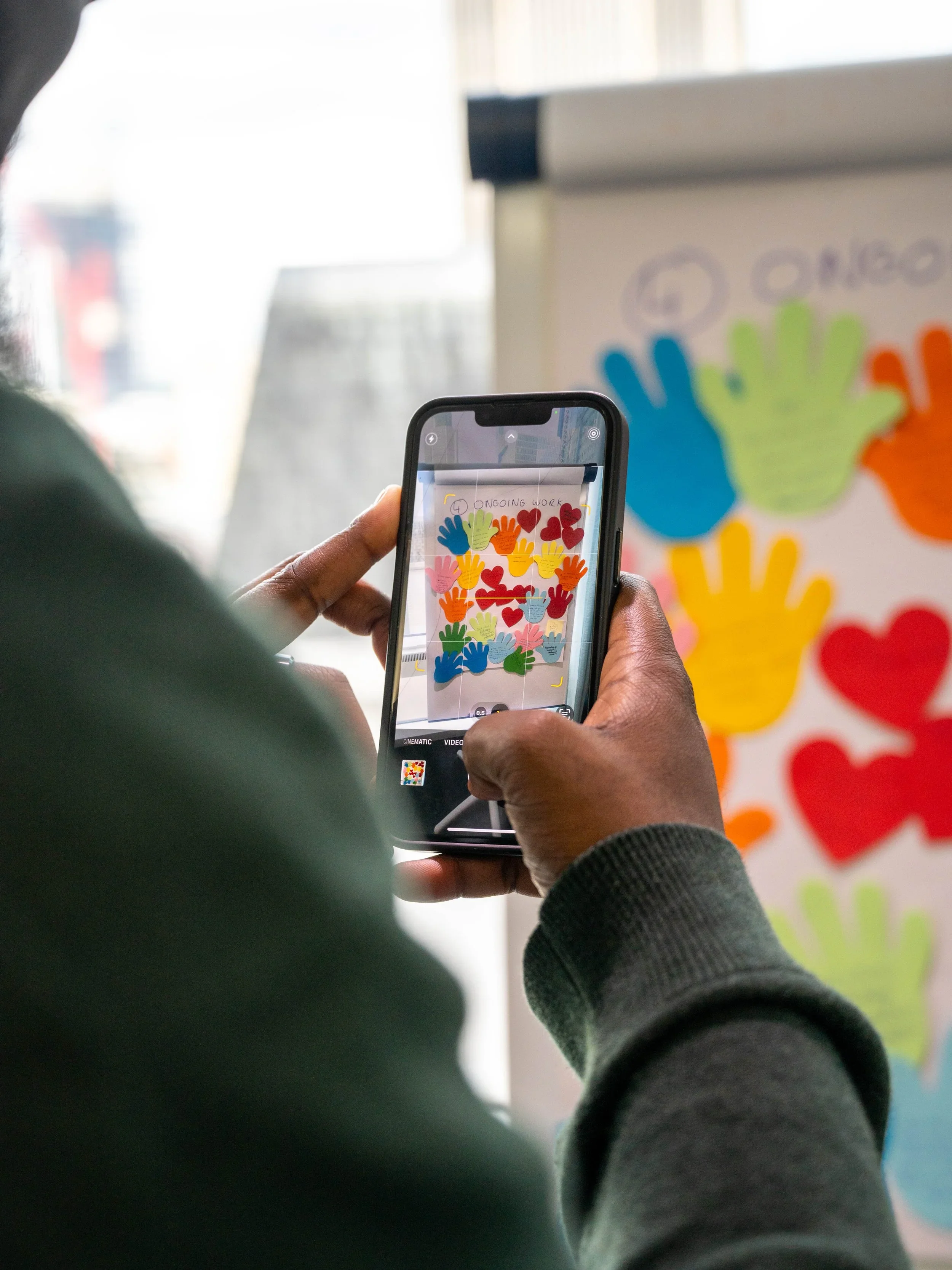 Photograph shows a person using a smartphone to capture a colorful community artwork display. Phone screen and poster feature overlapping blue, green, yellow, orange, and red handprints, hearts, and small handwritten messages.