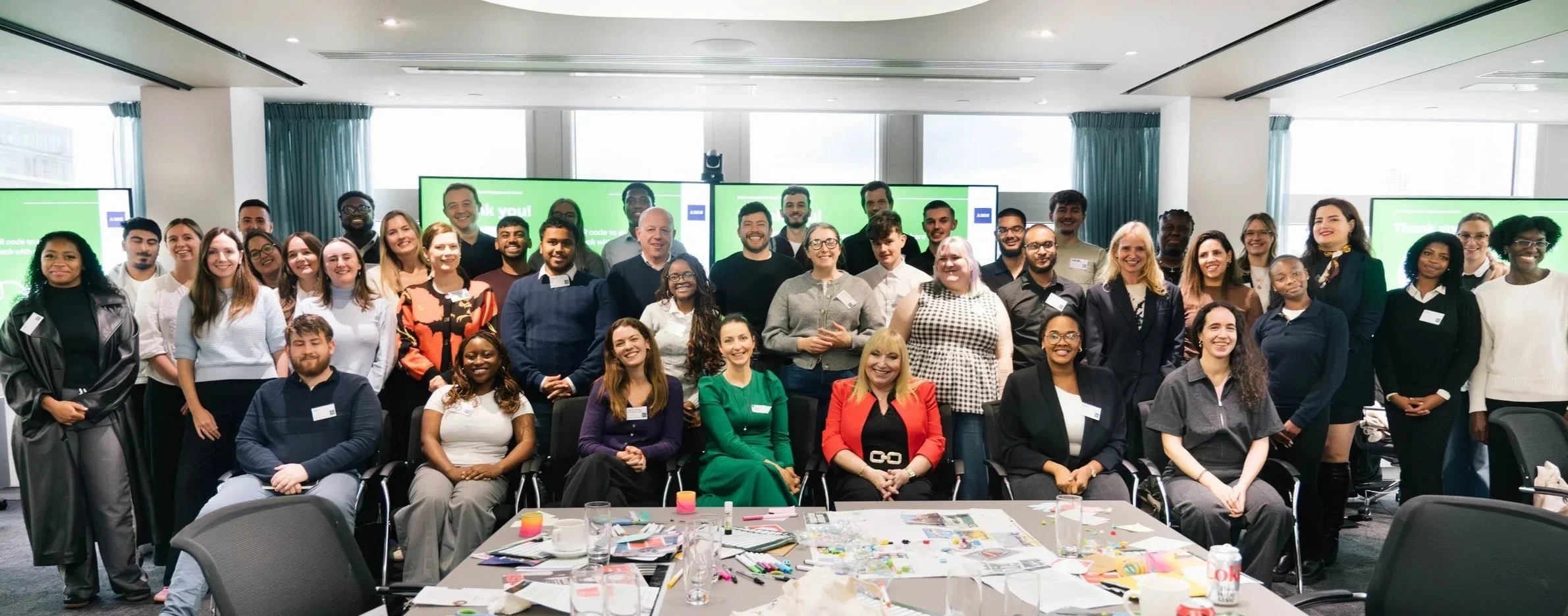 group photograph shows a diverse group of adults gathered for a collaborative workshop event in a bright conference room. Participants pose behind tables covered with colourful papers, markers, drinks and activity material with windows & curtains.