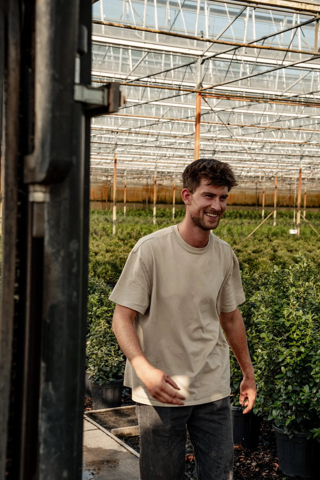A young man with brown hair and a beard smiling while working in a greenhouse among potted plants.