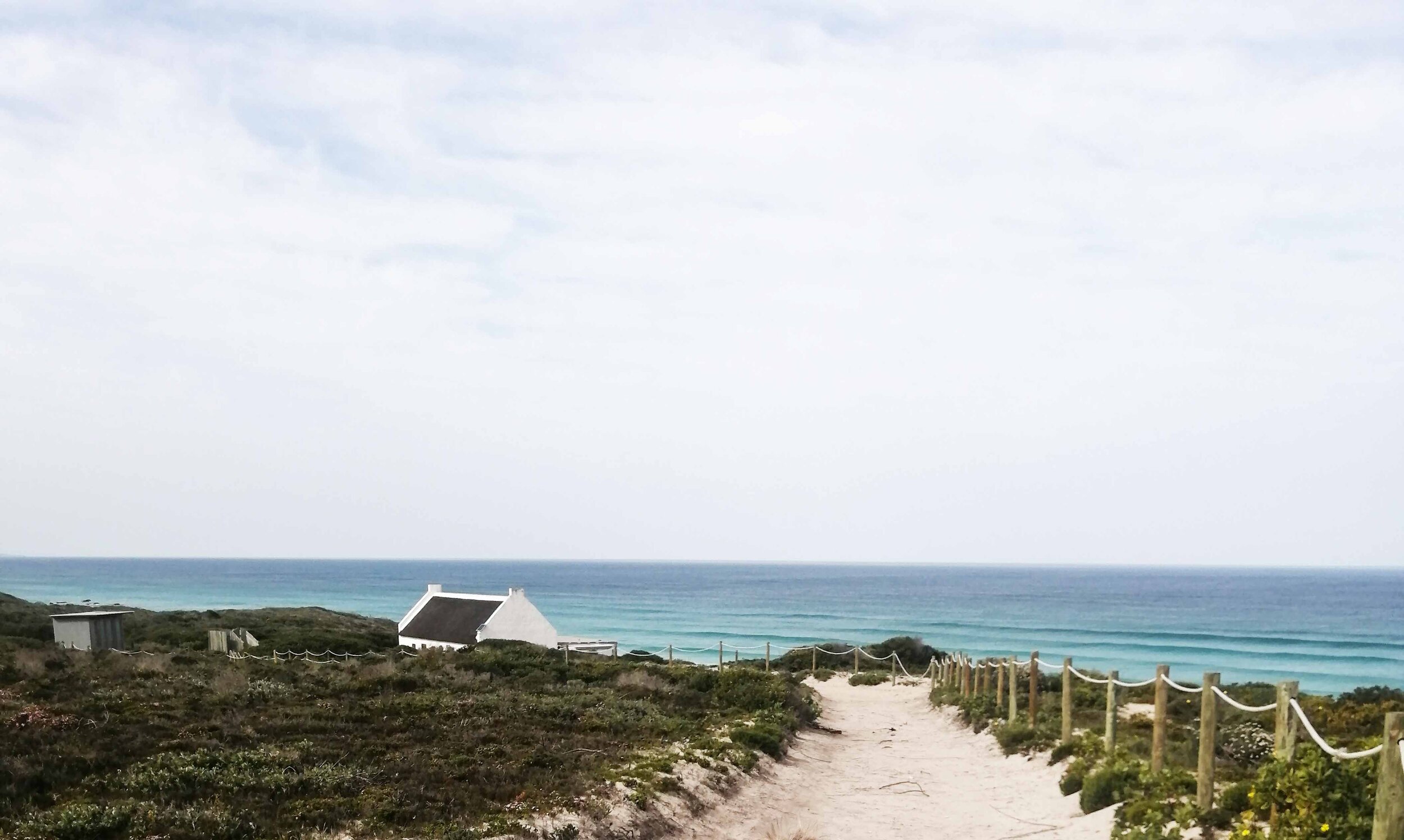 A coastal scene with a sandy pathway leading through vegetation to the beach, with small white buildings and a fence lining the path, overlooking the ocean under a cloudy sky.