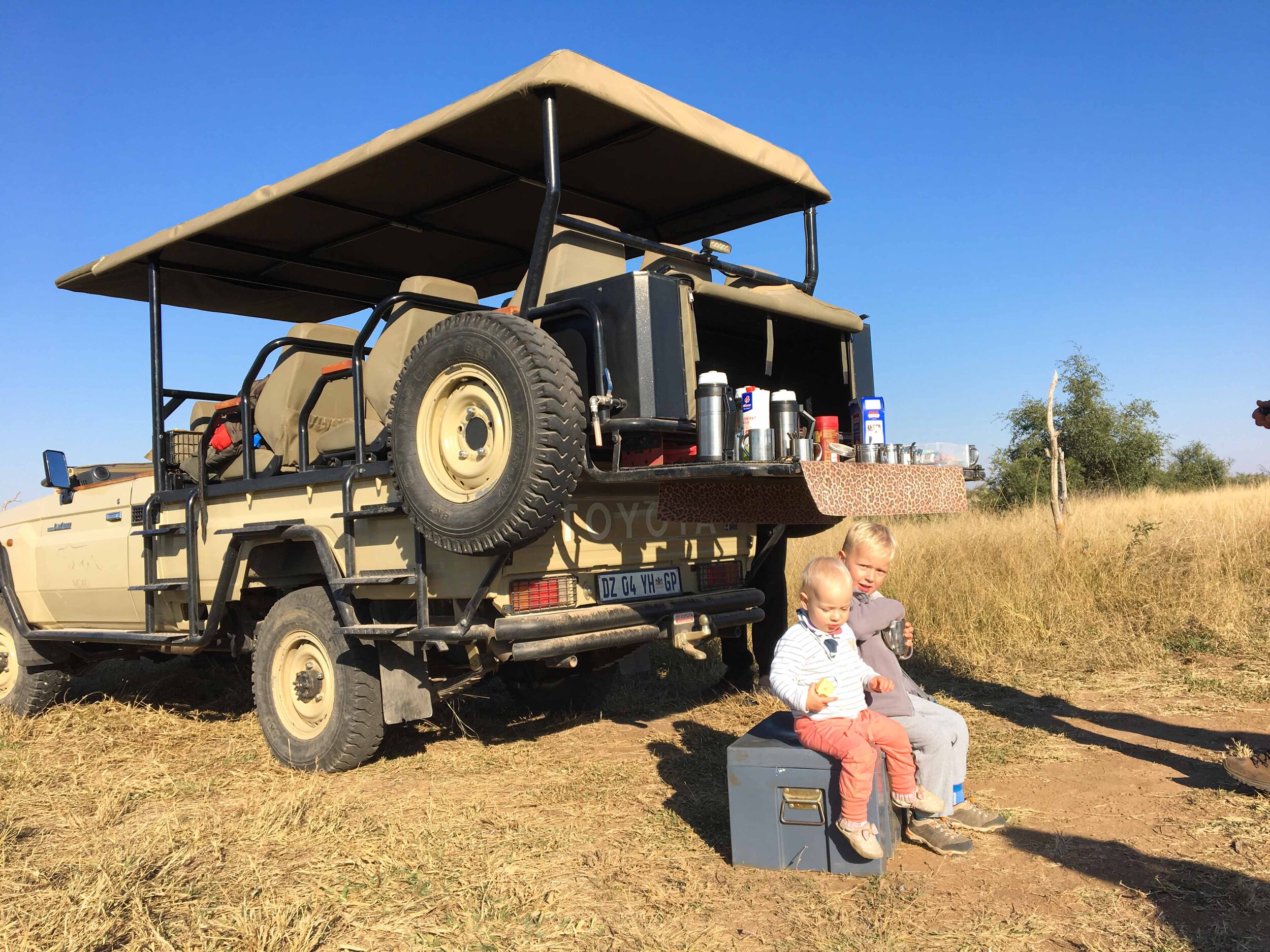 Two children sitting on a small gray bench beside a safari-style vehicle outdoors in a grassy field, with a clear blue sky and a few trees in the background.