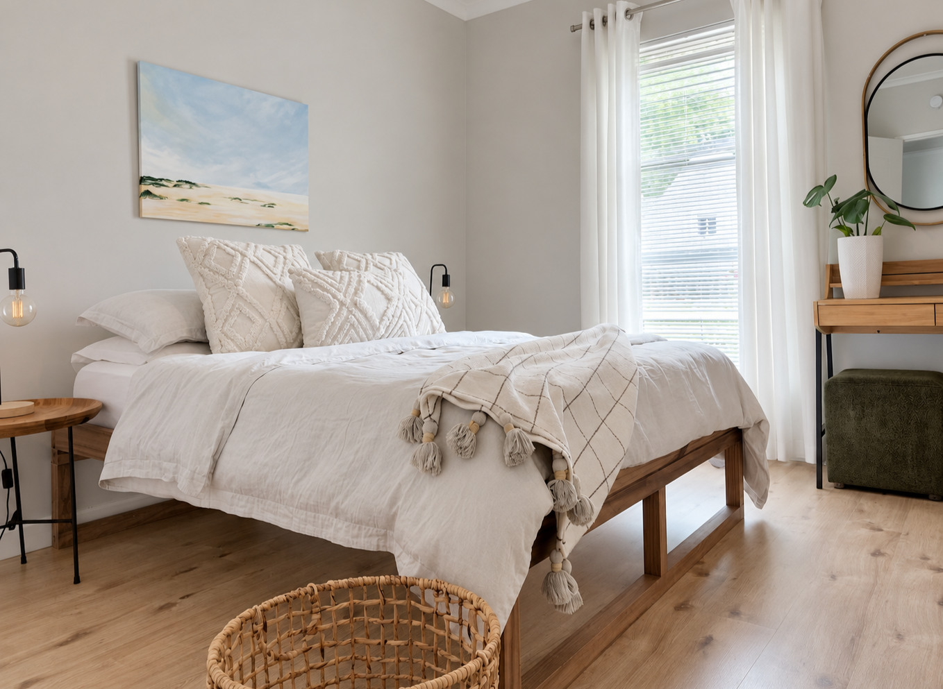 A cozy bedroom with a bed topped with white and cream textured pillows and a blanket, two hanging bedside lamps, a painting of a beach landscape above the bed, a window with white curtains, a wooden vanity with a mirror and a plant, and a woven basket on the wooden floor.