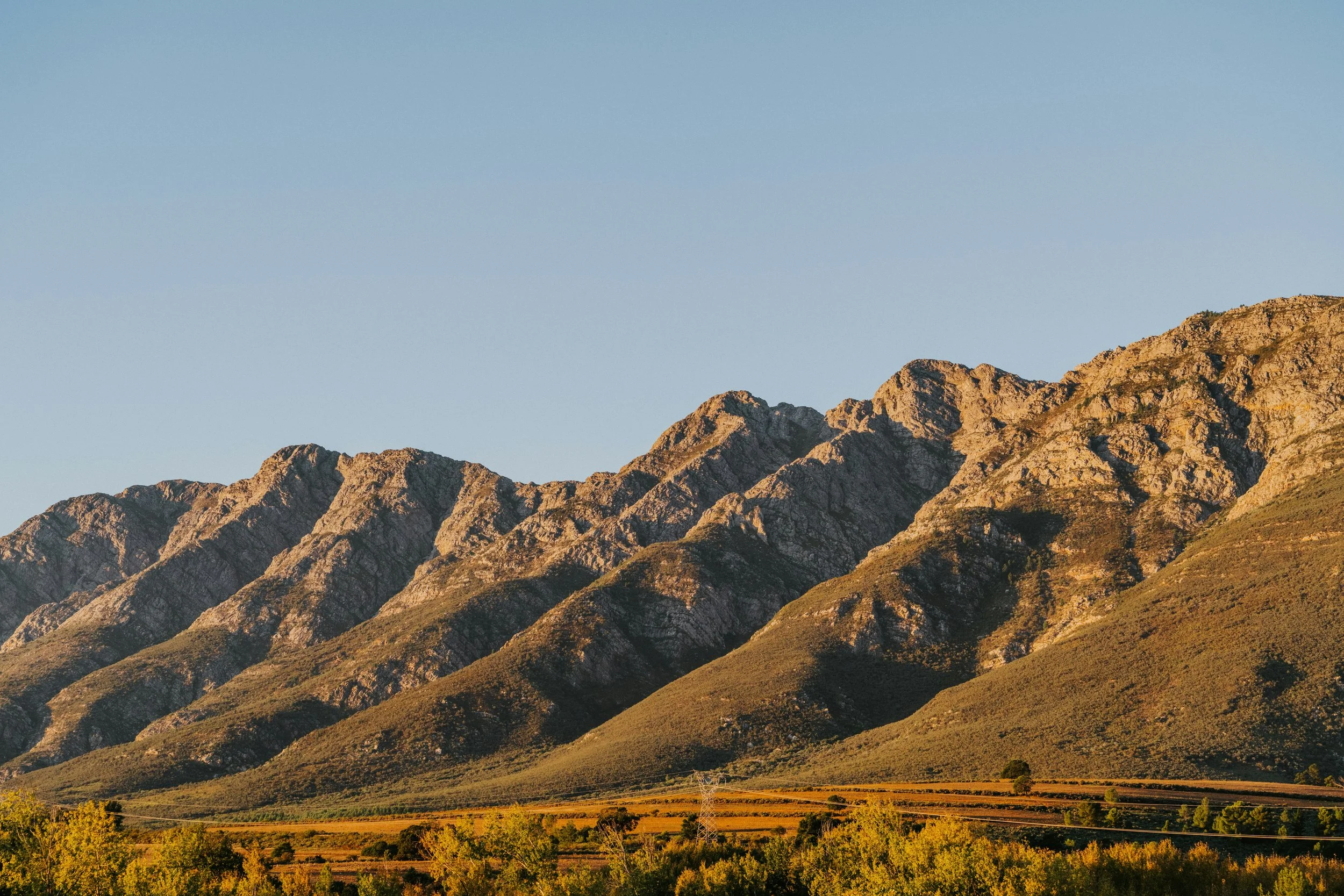 Sunlit mountain range with green and brown vegetation, clear blue sky above, and a power line in the foreground.