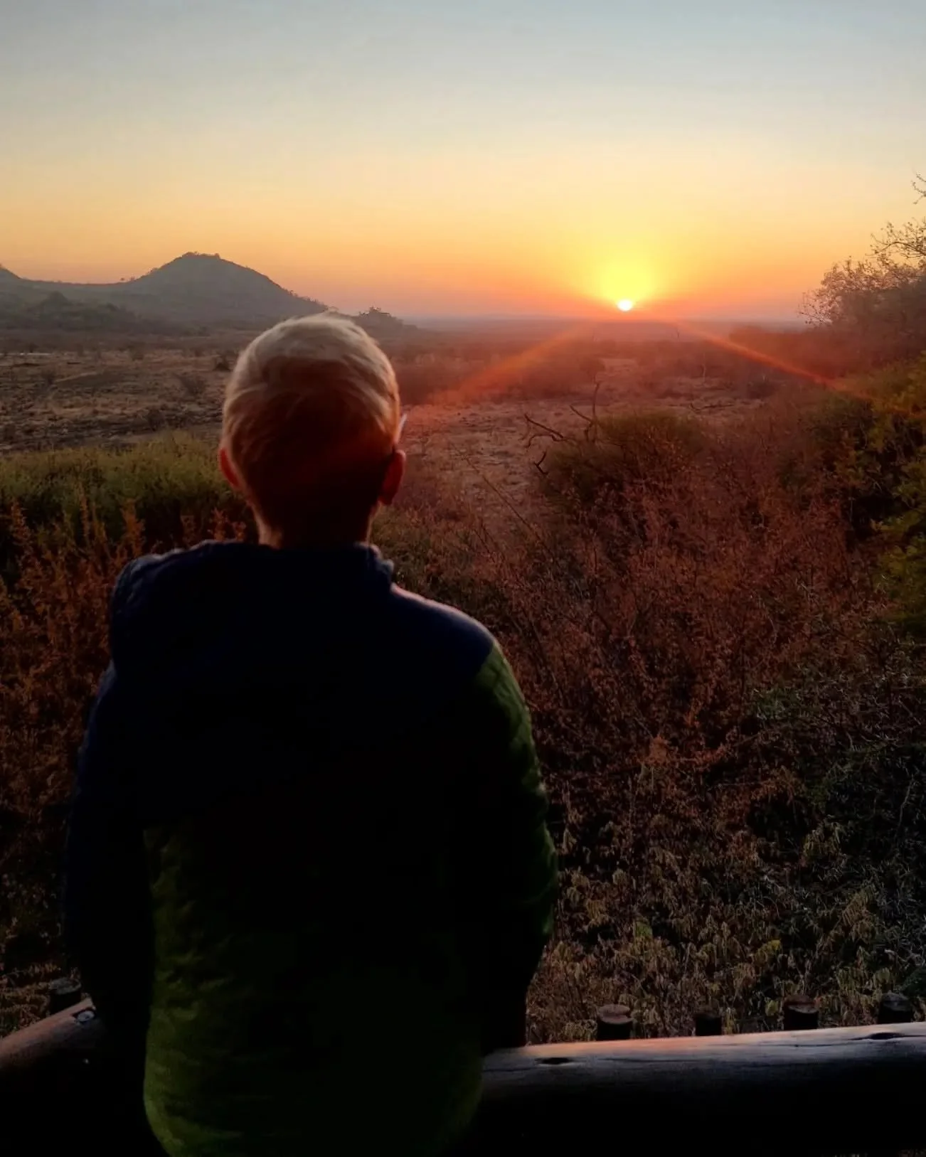 A person with short blonde hair, wearing a dark jacket, views a sunset over a landscape with hills and dry shrubs.