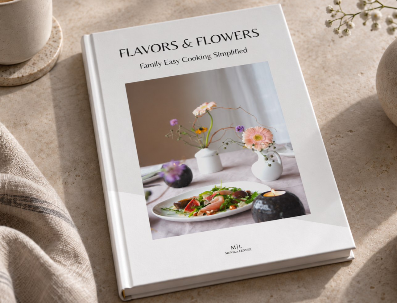 A cookbook titled "Flavors & Flowers: Family Easy Cooking Simplified" resting on a beige surface, featuring a cover photo of a plate of salad with various vegetables and garnishes, surrounded by vases with flowers and candles.