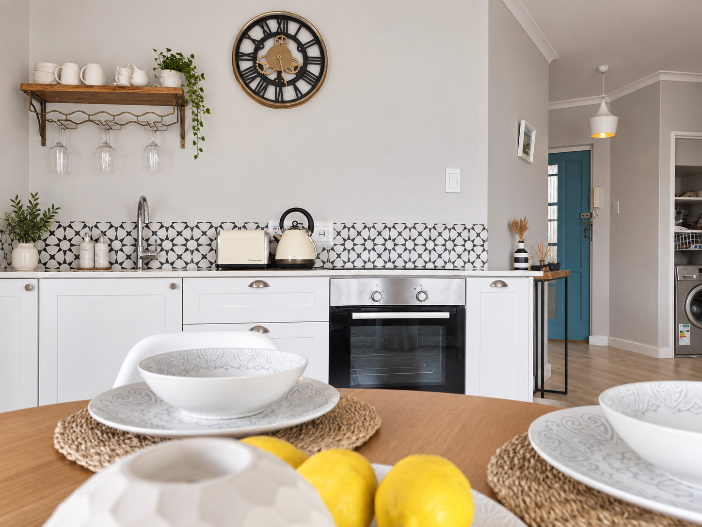 A modern kitchen with white cabinets, a geometric black and white backsplash, a black oven, and a countertop with a toaster and kettle. A wooden shelf with mugs and a plant is above the sink, and a large clock hangs on the wall. The dining table in front has white bowls, lemon-shaped decorations, woven placemats, and is set for meals. To the right, there's a blue door and some additional kitchen storage units.