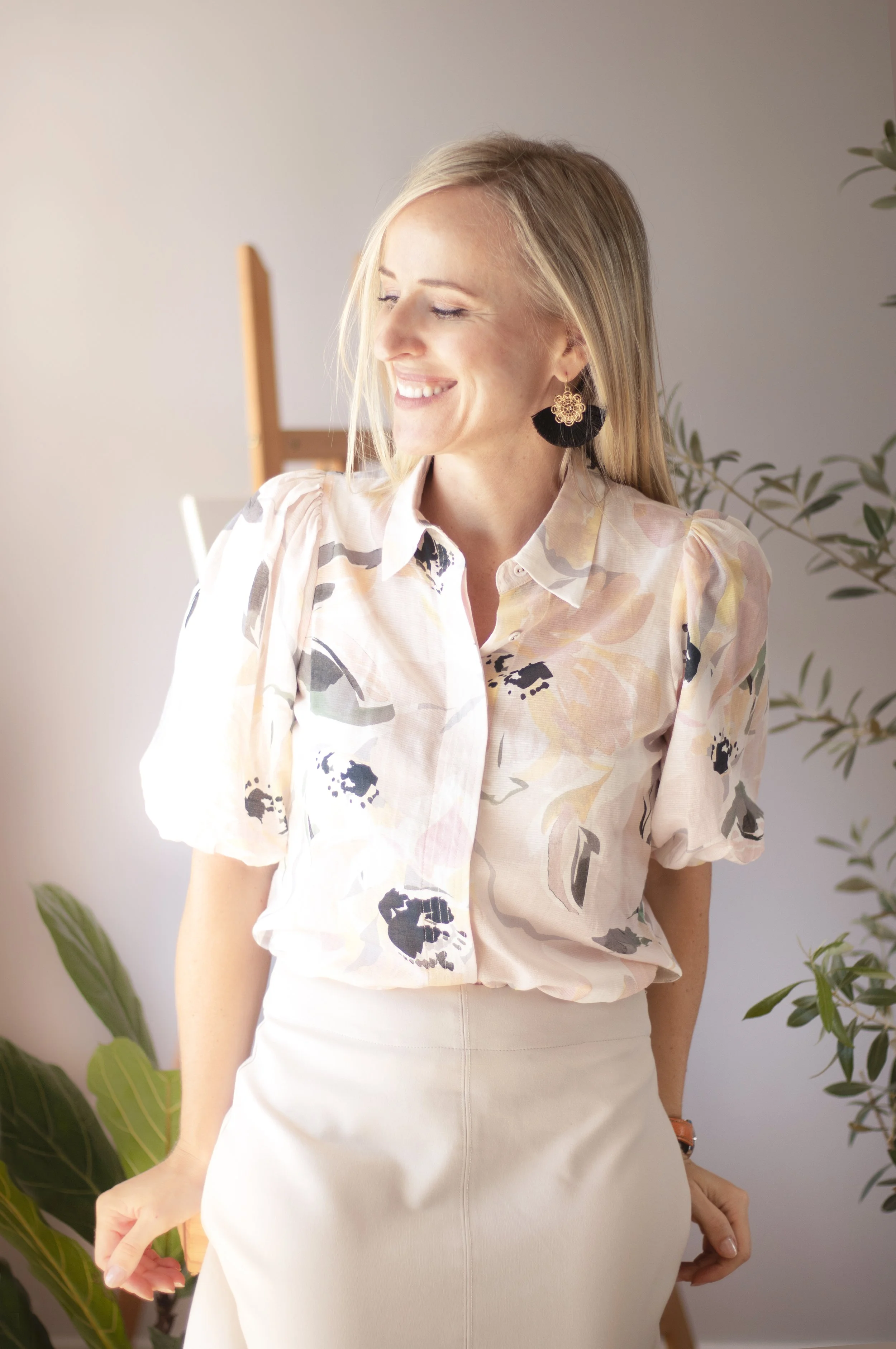 A woman with blonde hair wearing a floral blouse and cream-colored skirt, smiling with closed eyes, standing indoors near green plants.