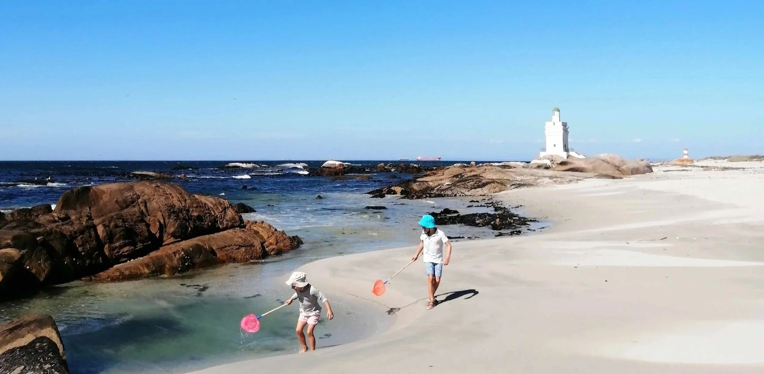 Two children, a boy and a girl, playing with nets on a sandy beach near rocks, with a lighthouse and the ocean in the background under a clear blue sky.