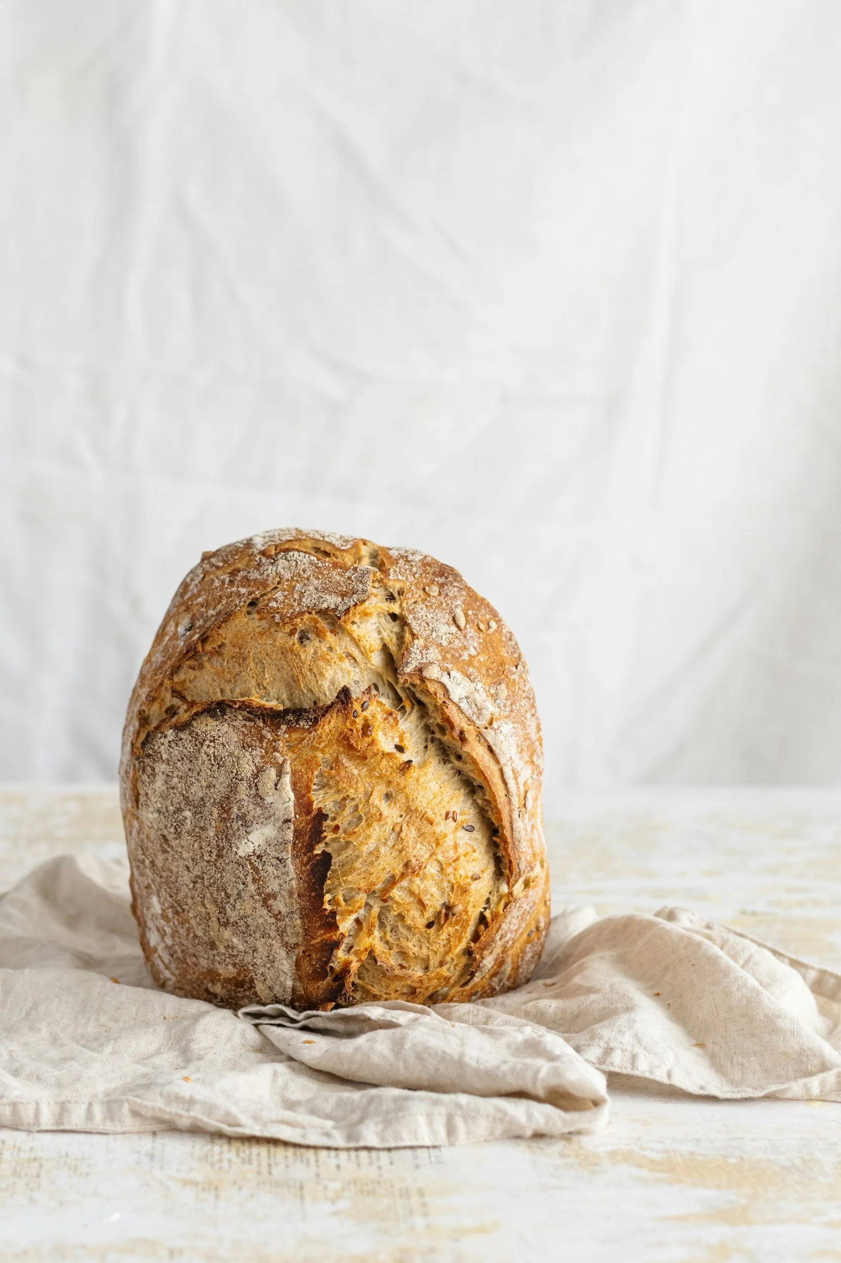 Krustiges, ovales Brot mit Mehl bestäubt, auf einem weißen, leicht gewellten Tuch liegend auf einem hellen Holztisch, gegen einen weißen Hintergrund