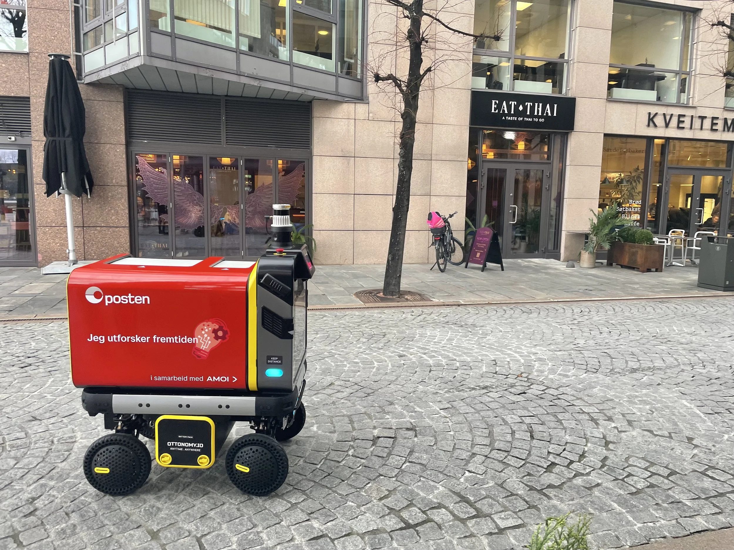 A small autonomous delivery robot on a city sidewalk in front of a bakery and cafe. The robot has a red and black body with a yellow section, and displays the logo and text in Norwegian. A bike is parked nearby, and a tree lines the street. In the background, there is a modern building with large glass windows, one of which features a pink angel wings decal.