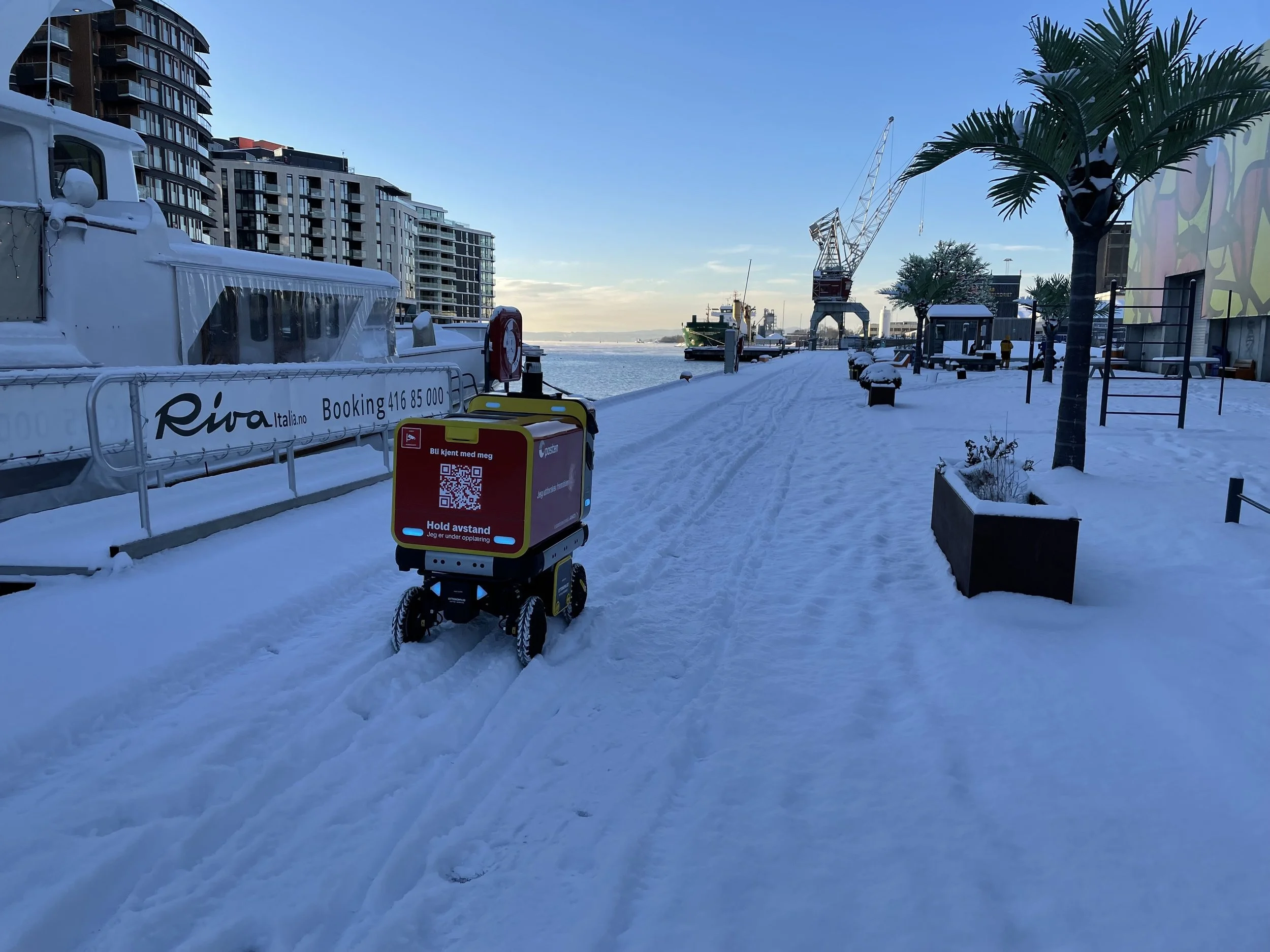 Snow-covered walkway along a waterfront with modern buildings, palm trees, a ship, industrial crane, and a robotic delivery device in the foreground.