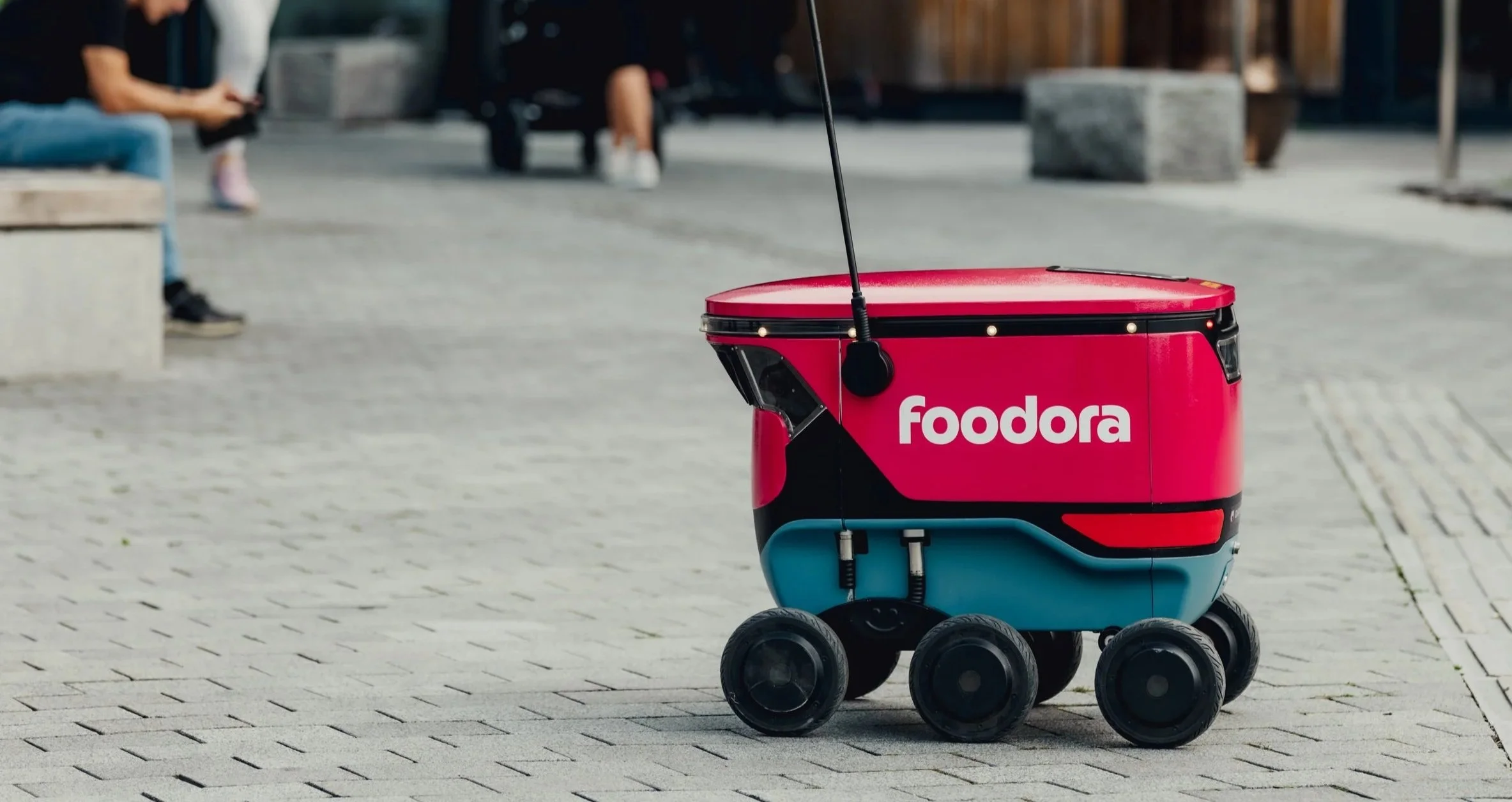 A Foodora branded delivery robot on a city sidewalk with blurred pedestrians in the background.