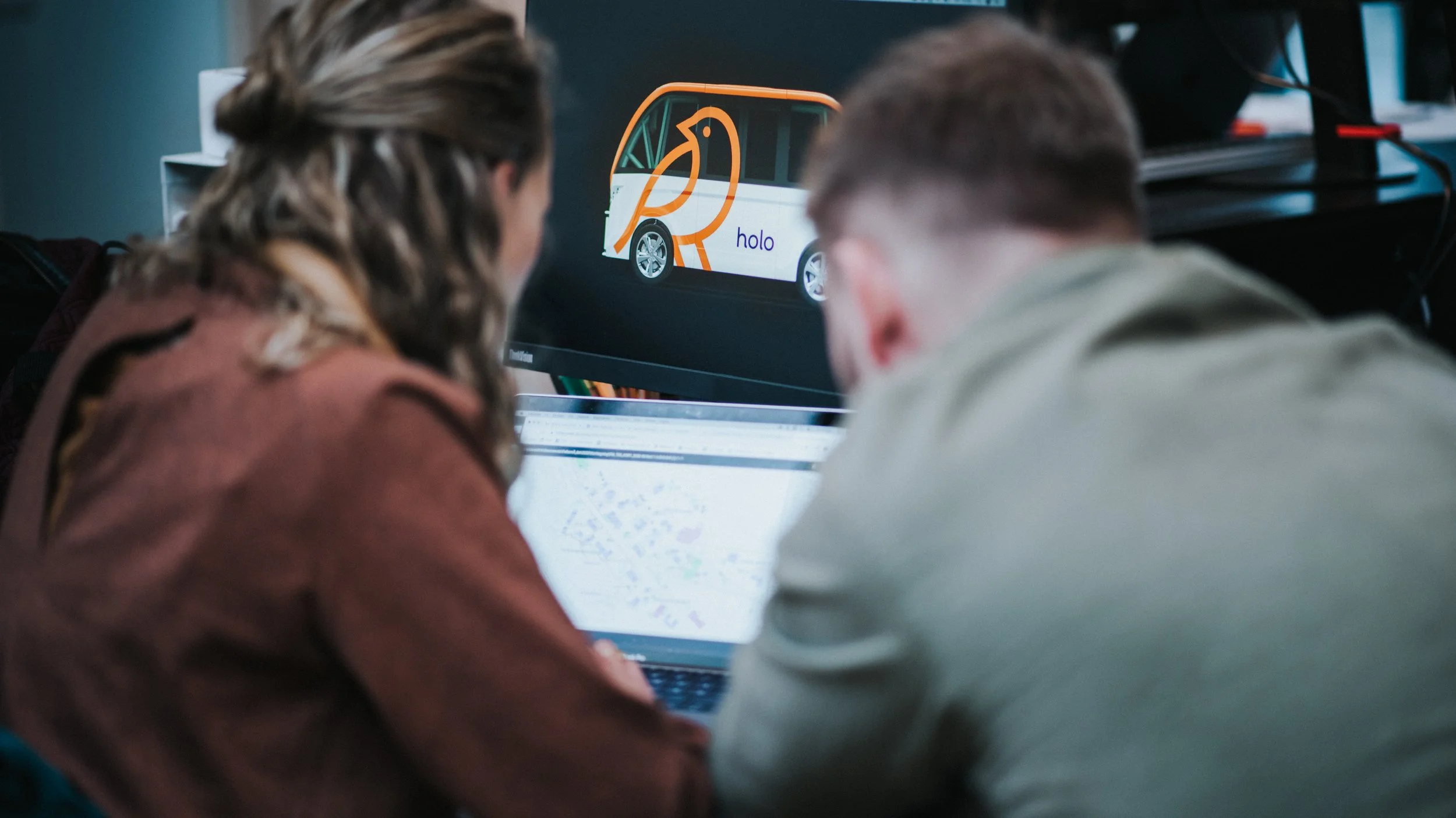 Two people sitting at a desk looking at a computer screen displaying a vehicle with a logo and the word 'holo' on it.