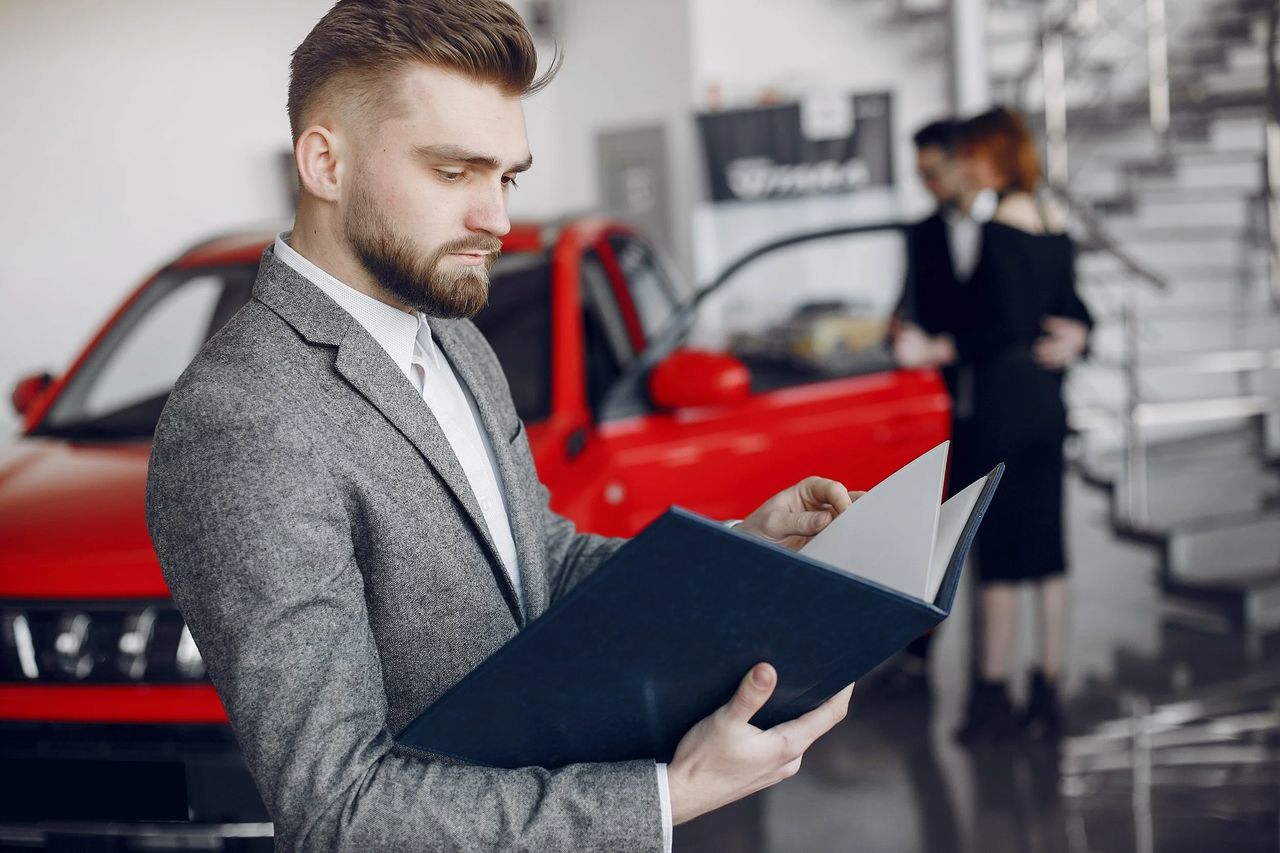 A man in a gray blazer is standing in a car dealership, looking at a folder. In the background, a woman with earphones and a black dress is near a red car.