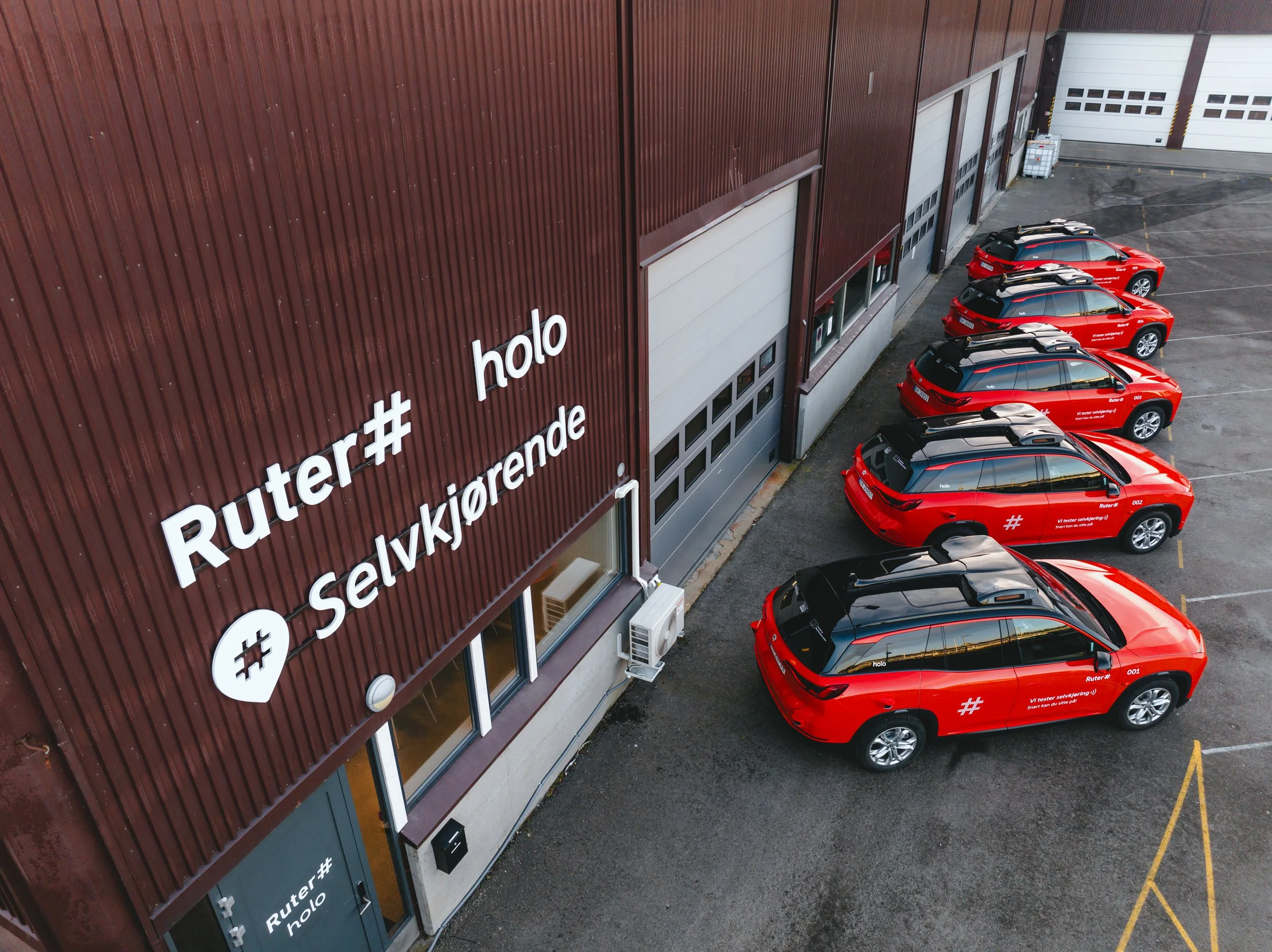 Red cars parked in a parking lot next to a large industrial building with a brown exterior wall. The building has multiple white garage doors and signs with hashtags and Norwegian text.