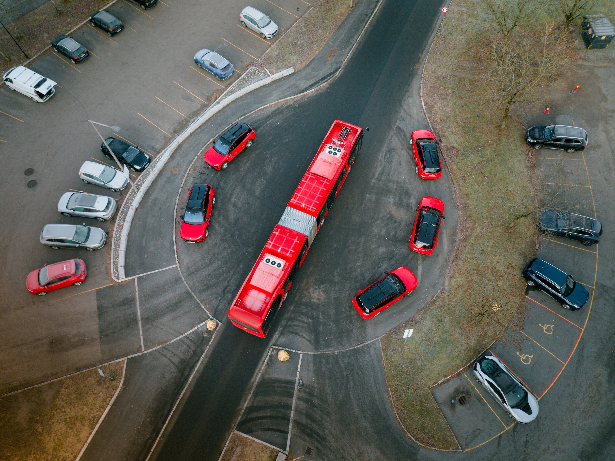 An aerial view of a parking lot with several parked cars, a street, some trees, and a red bus on the road.