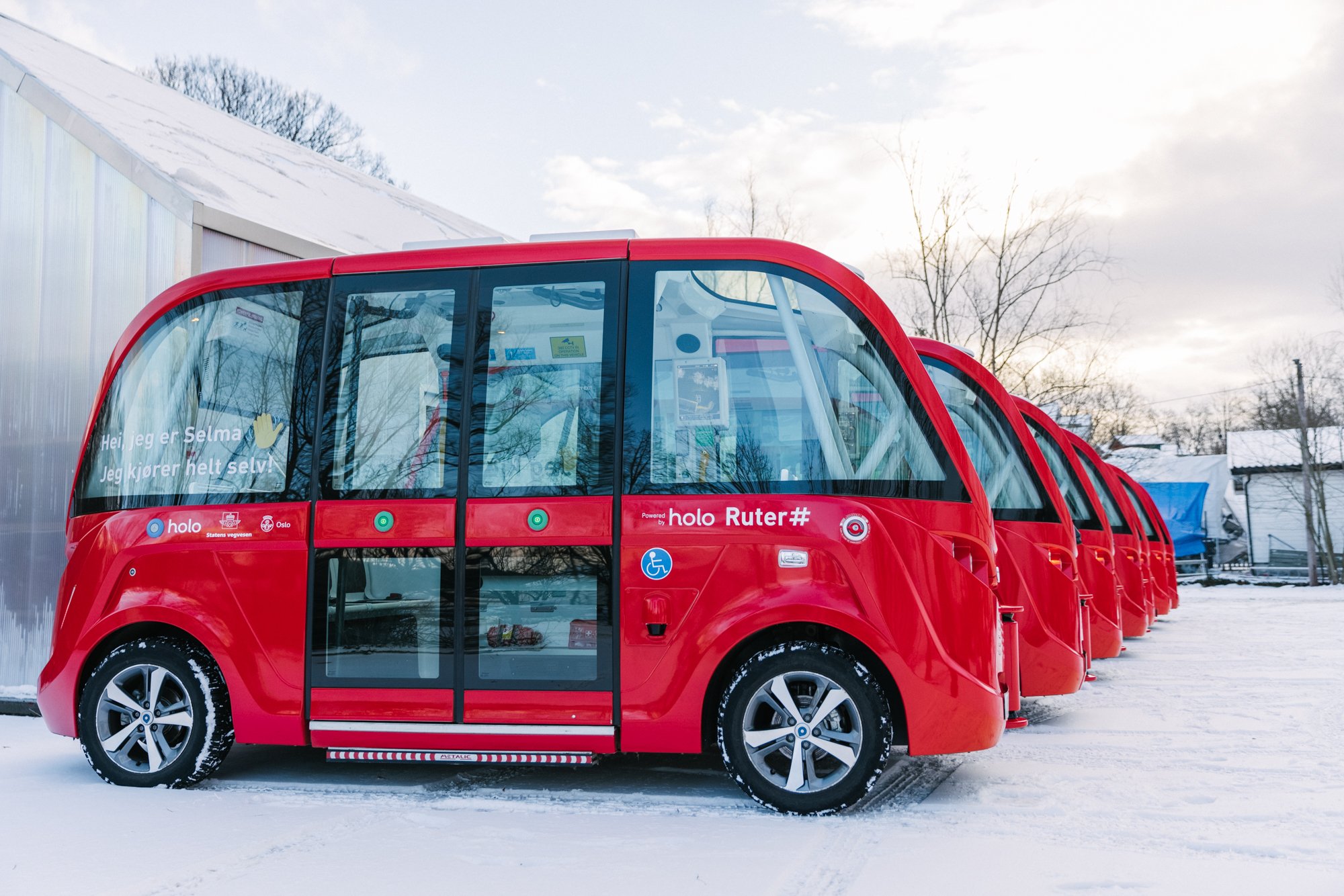 A row of red autonomous shuttle vehicles parked on snow-covered ground with a building and bare trees in the background. The shuttle has large windows and a Norwegian text on the side.