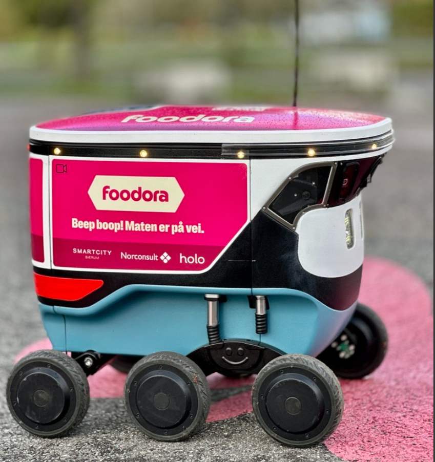 Autonomous food delivery robot with a pink and black exterior on a sidewalk.