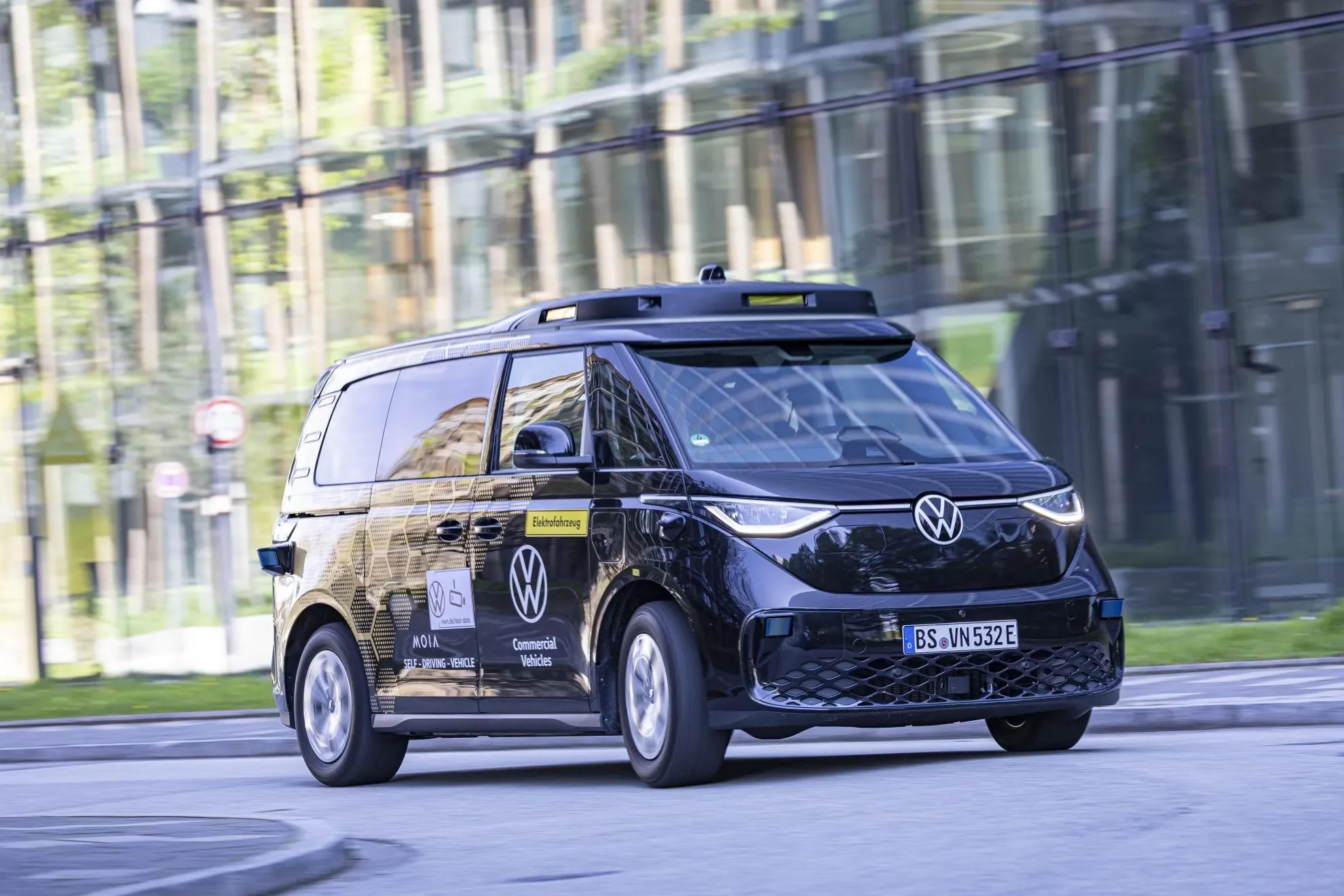 Black Volkswagen self-driving electric van with yellow and black markings, driving on city street with modern glass building in background.