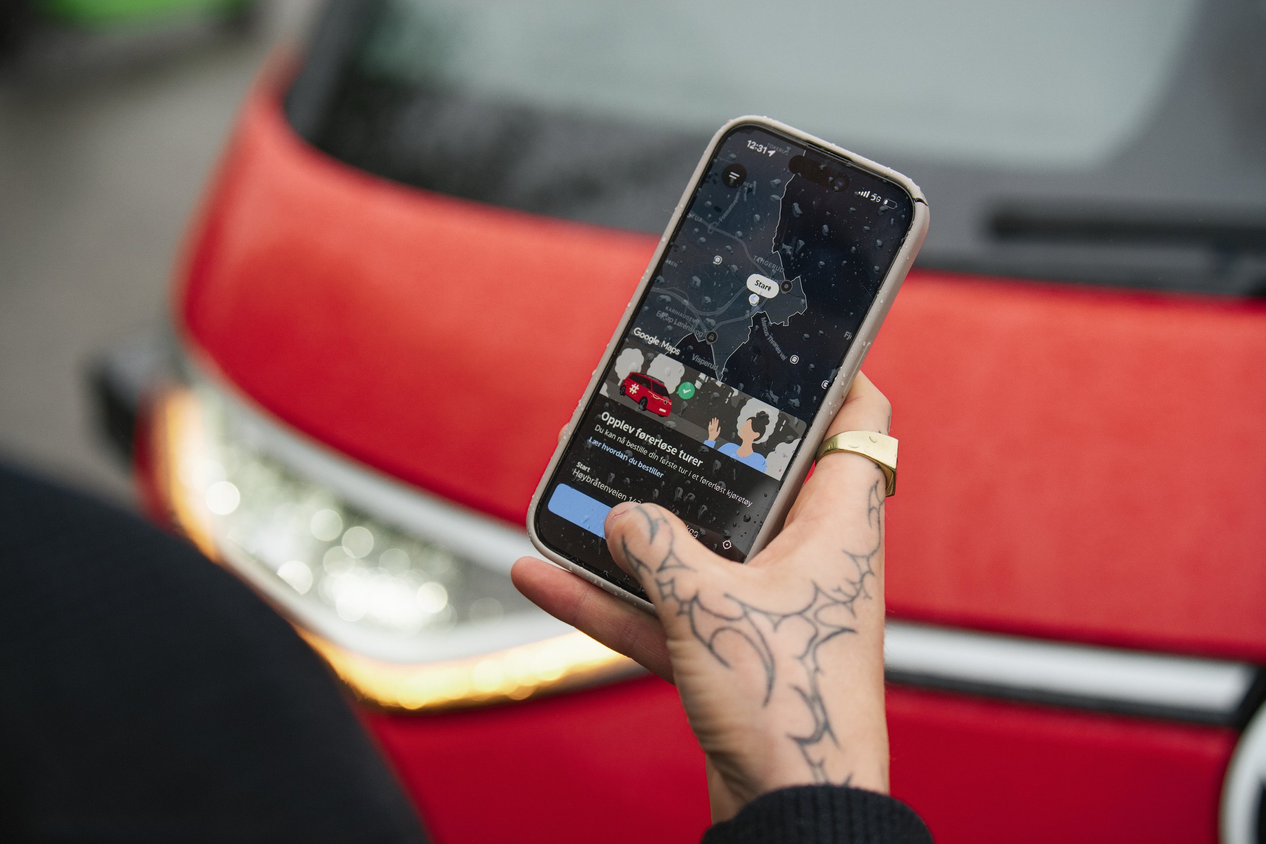 Person with tattoos on hand holding a smartphone displaying Google Maps app; red vehicle in background with rain droplets on the phone screen.