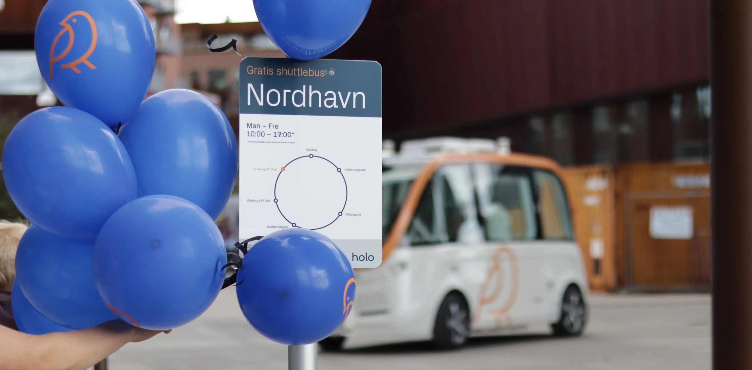 A person holding blue balloons with a public transit schedule sign for Nordhavn station in the background.