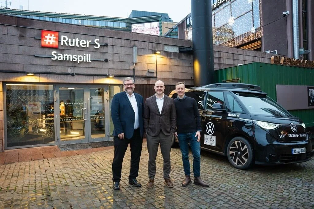 Three men standing in front of a building with a Volkswagen self-driving vehicle parked beside them. The building has a sign that reads '# Ruter S Samspill.' The men are dressed in business casual attire, and the setting appears to be in an urban area.