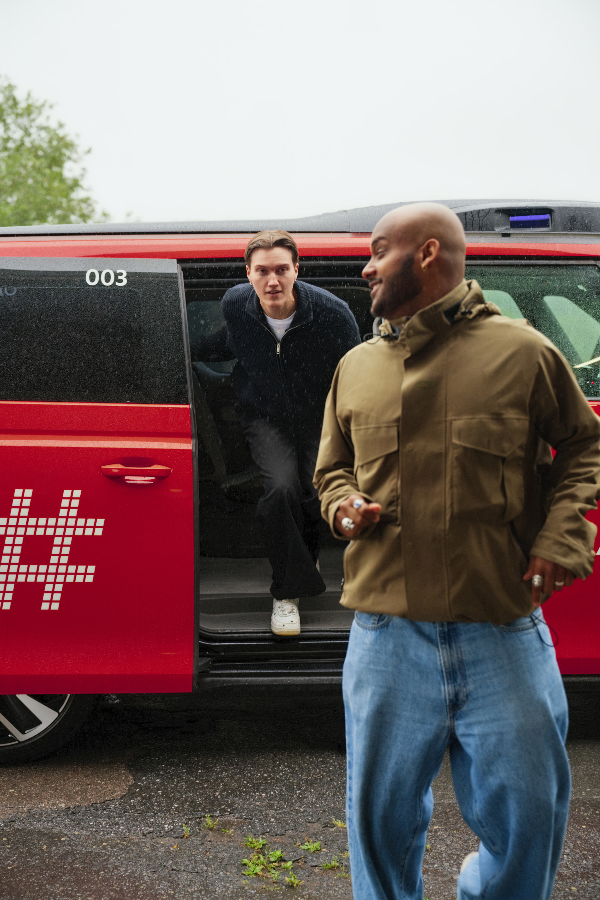 Two men, one with a shaved head and the other with brown hair, engaging with each other outdoors near a red autonomous Holo vehicle, with one leaning out of the vehicle window and the other standing nearby.
