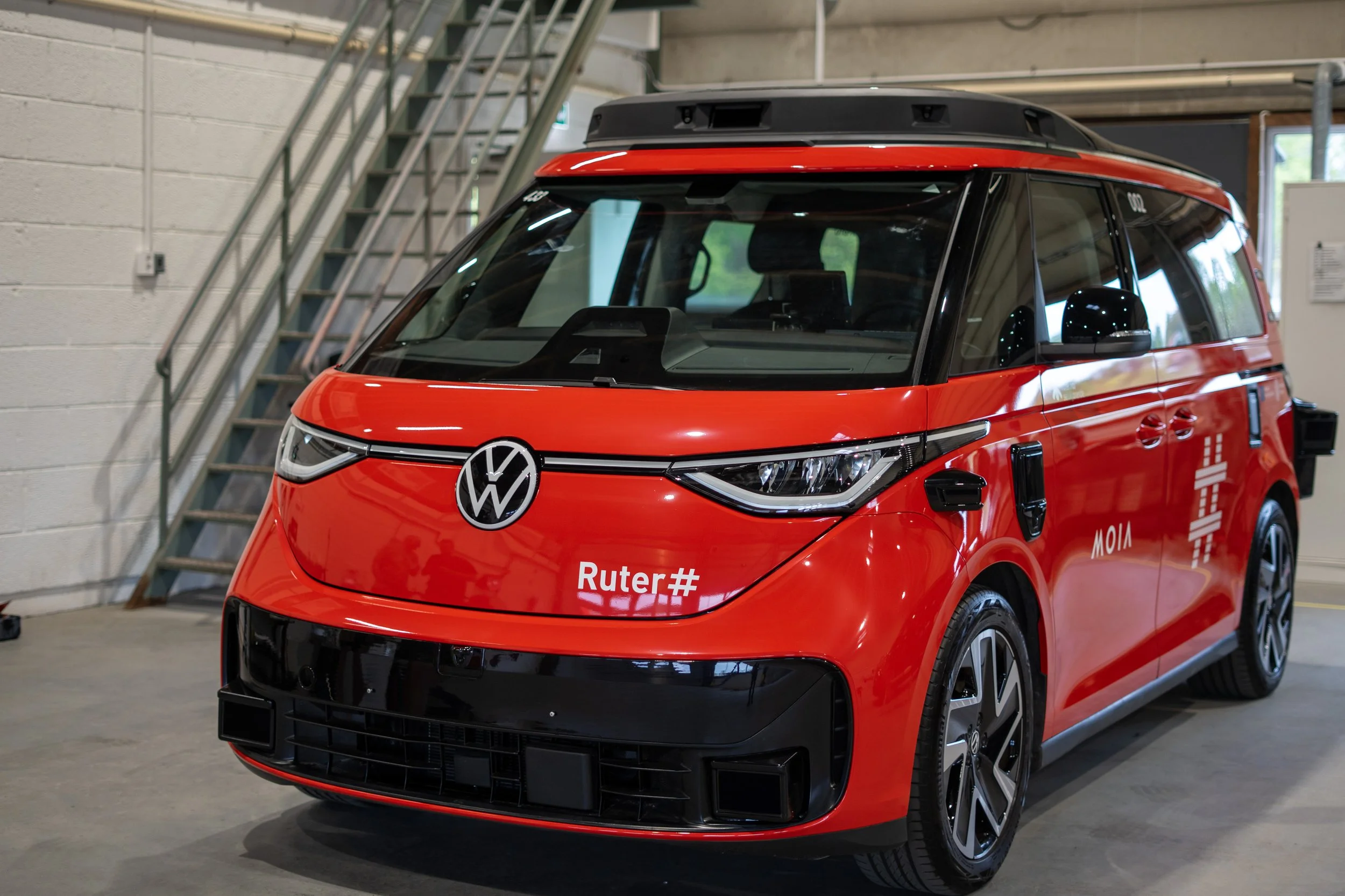 Red Volkswagen electric van parked indoors with large front windshield, side mirrors, and black roof, featuring text in Norwegian on the side that reads 'Ruter' and 'MOI'.