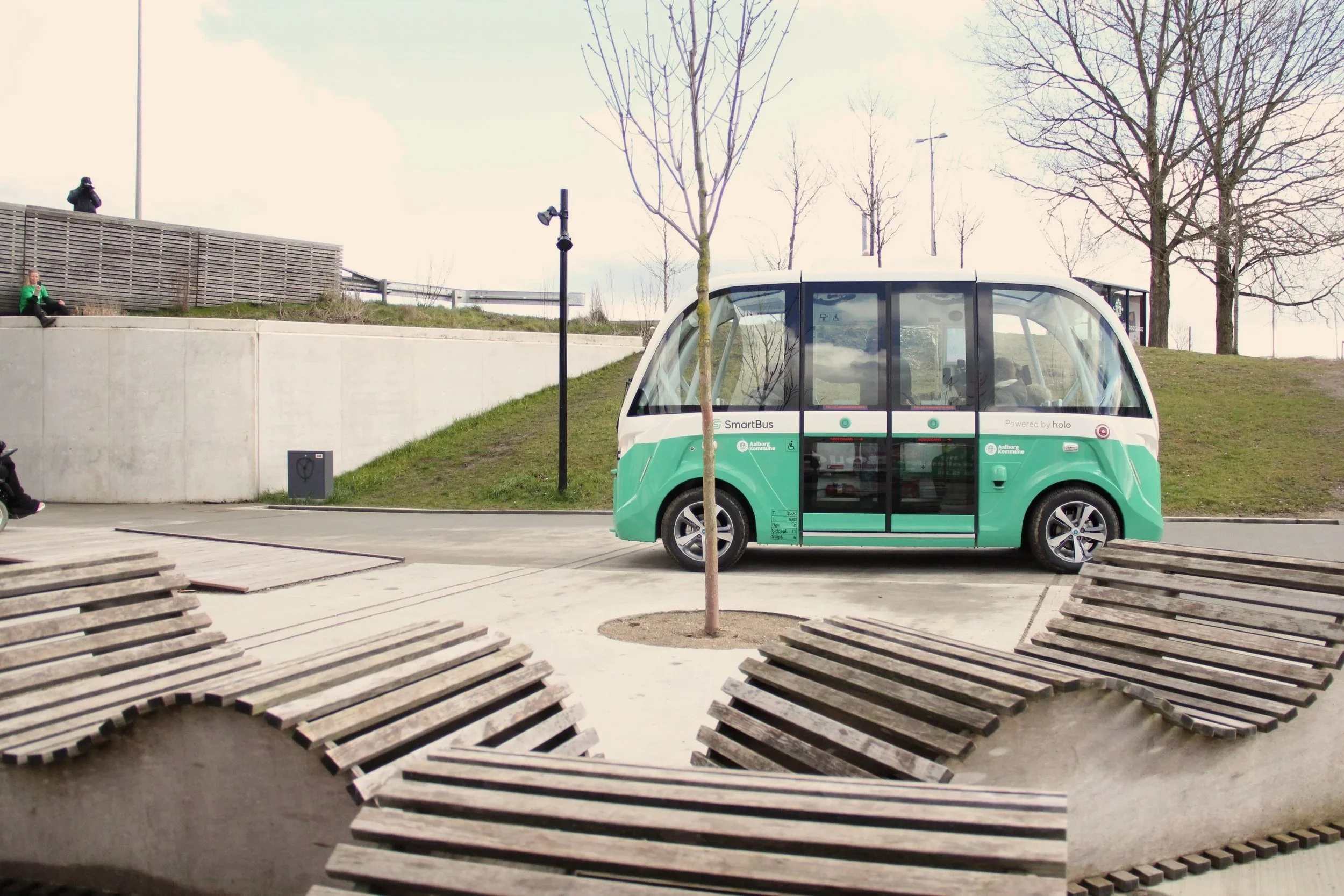An electric shuttle vehicle parked near a sidewalk with wooden benches around a small tree, in an outdoor area with a grassy hill and leafless trees in the background.
