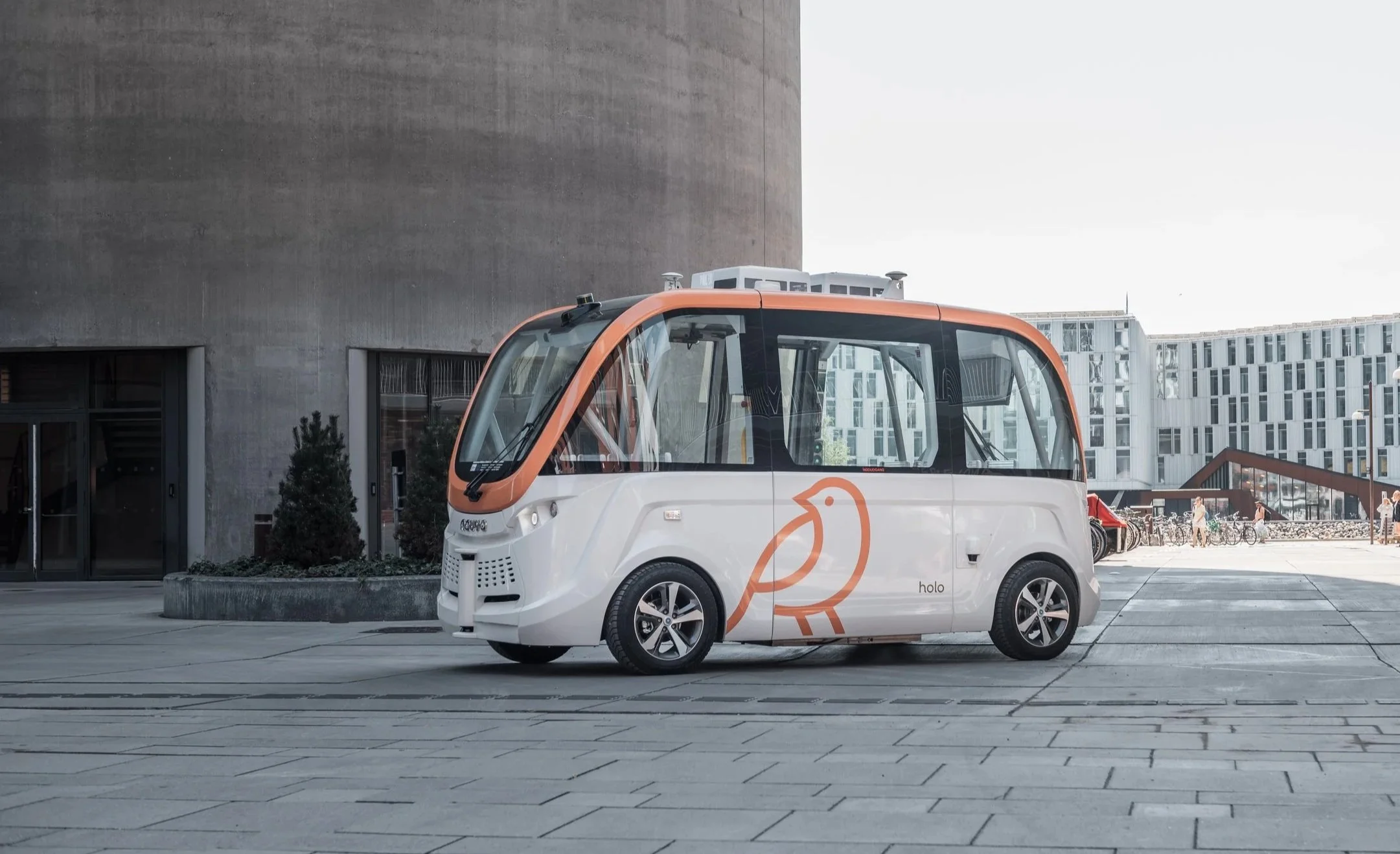 Electric delivery vehicle with a white and orange color scheme, featuring a stylized bird logo, parked in an urban area with modern buildings and bicycles in the background.