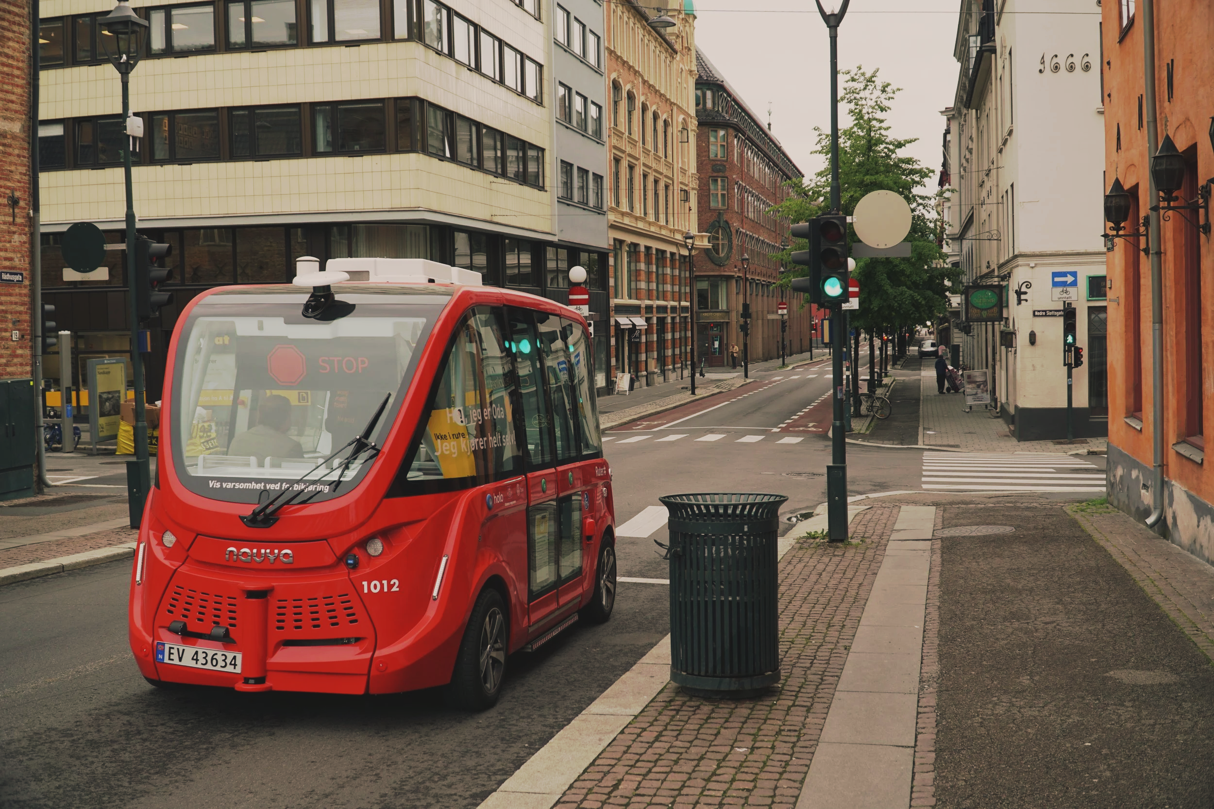 A red autonomous shuttle vehicle driving through a city street with modern buildings, streetlights, and traffic signals, under an overcast sky.