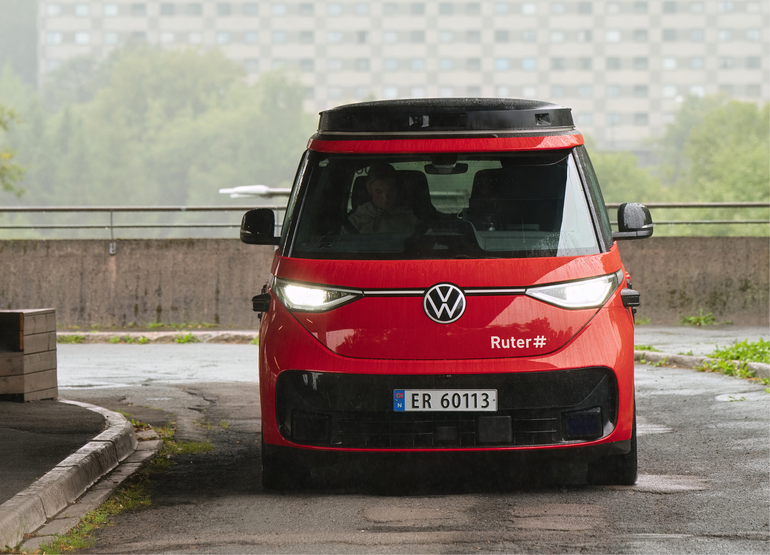 Red autonomous vehicle with Volkswagen logo, license plate ER 60113, and #Ruter hashtag, parked near a curb with trees and a multi-story building in the background.