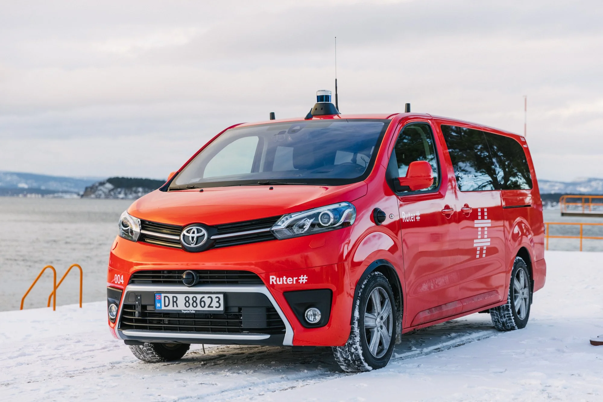 Red  autonomous Holo vehicle with roof-mounted sensors and antennas, parked on snow near a body of water with a rocky shoreline and trees in the background.