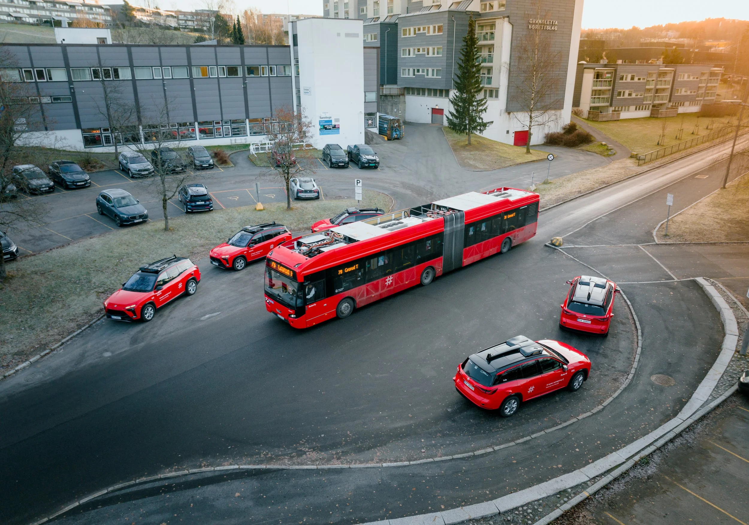 A city street scene with a red bus and red cars parked along the roadside, modern buildings in the background, and a partly cloudy sky with the sun setting.