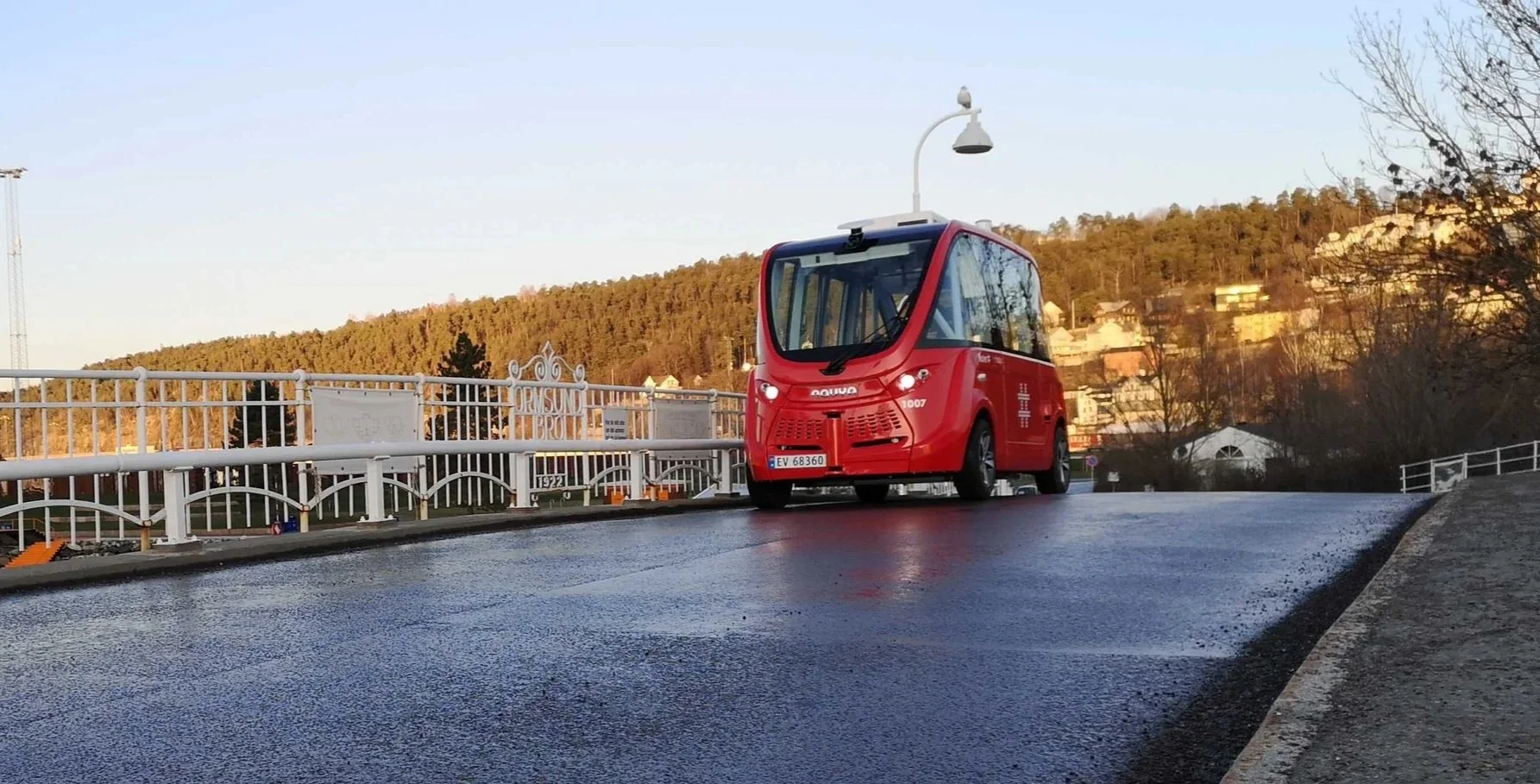 A small red autonomous electric vehicle driving on a wet road, with a white railing on the side and a streetlamp overhead, in a scenic area with houses and trees on a hill in the background.