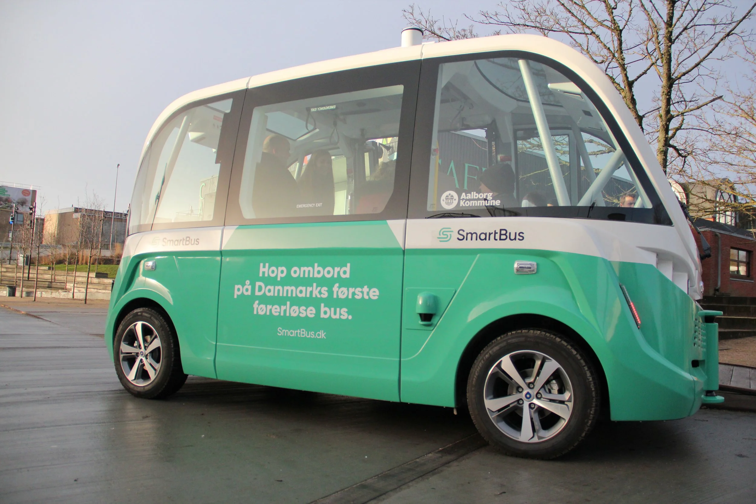 A small green and white autonomous bus labeled 'SmartBus' on a paved area with leafless trees and buildings in the background.