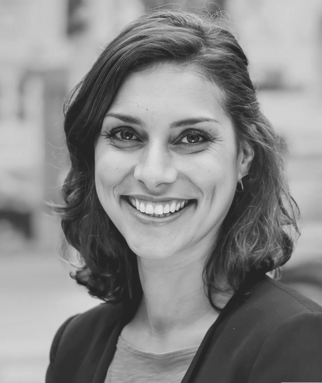 Black and white photograph of a smiling woman with shoulder-length hair, wearing a blazer and earrings, outdoors in an urban setting.