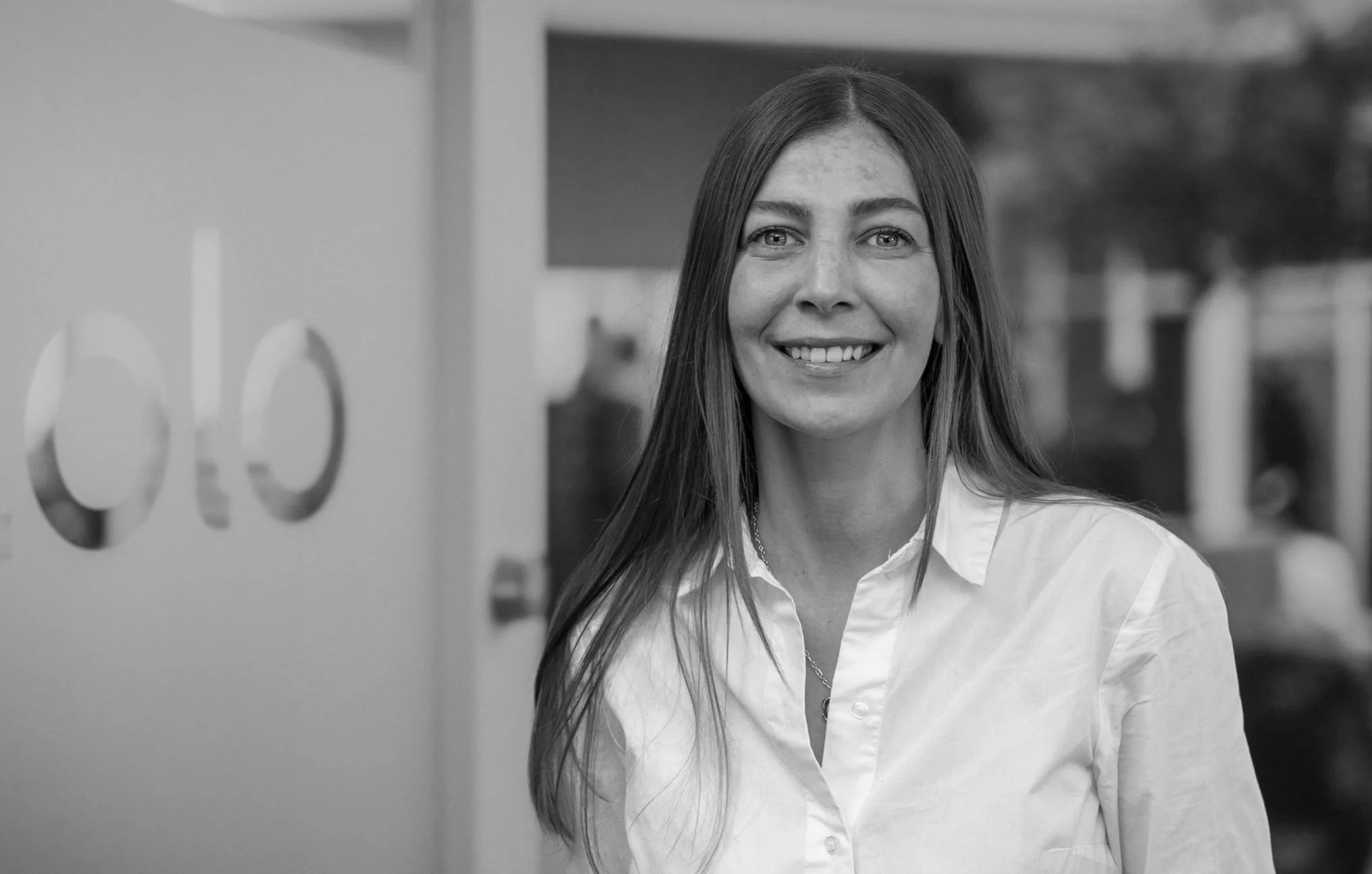 A smiling woman with long hair wearing a white shirt inside a building with abstract wall art.