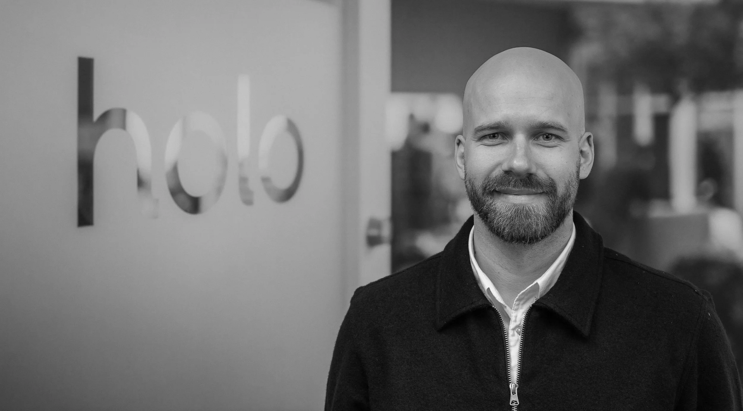 A man with a beard and a shaved head smiling outdoors near a glass wall with the partially visible word 'hope' on it.