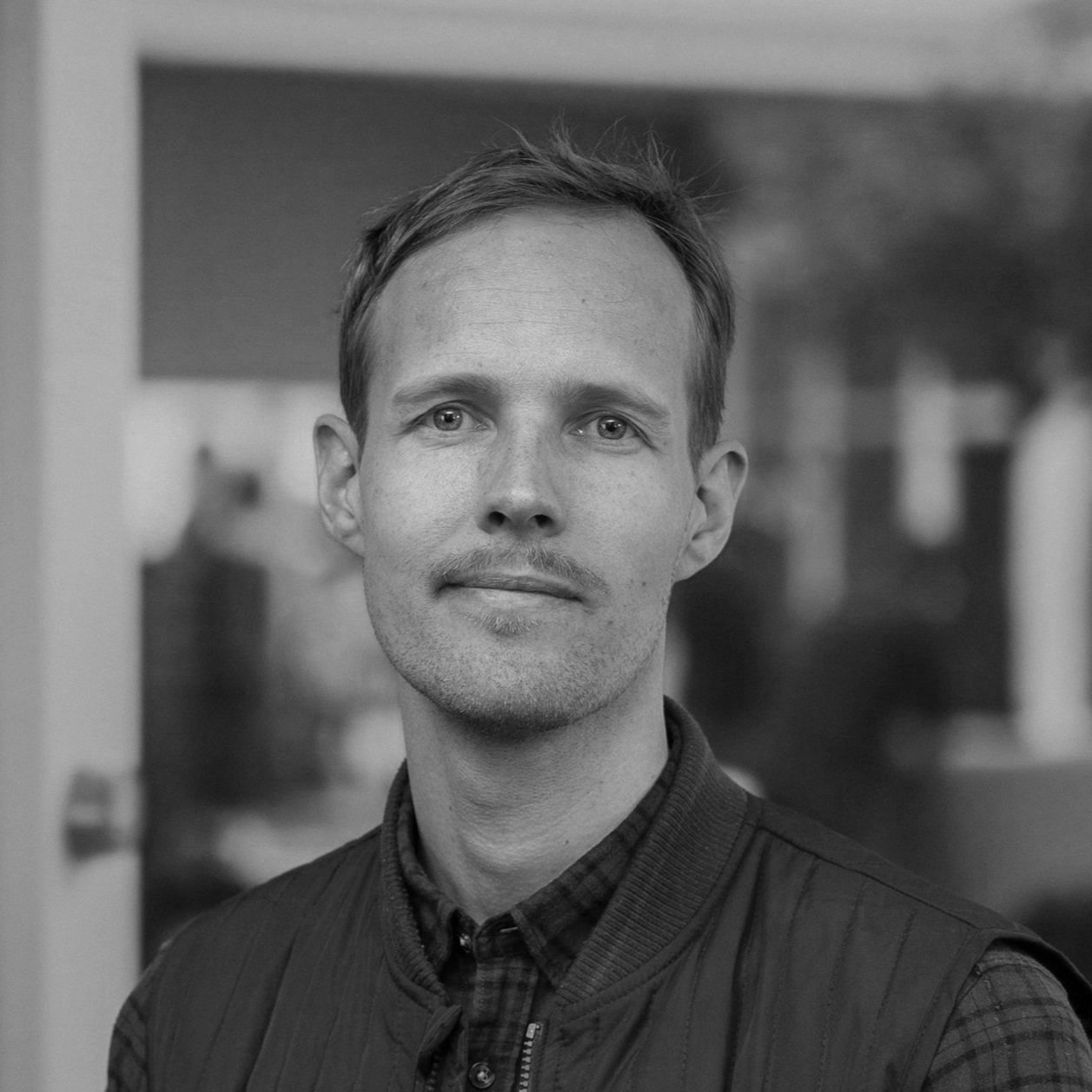 A black and white portrait of a man with short hair, light eyes, and a slight smile, wearing a collared shirt and vest, standing outdoors with blurred background elements.