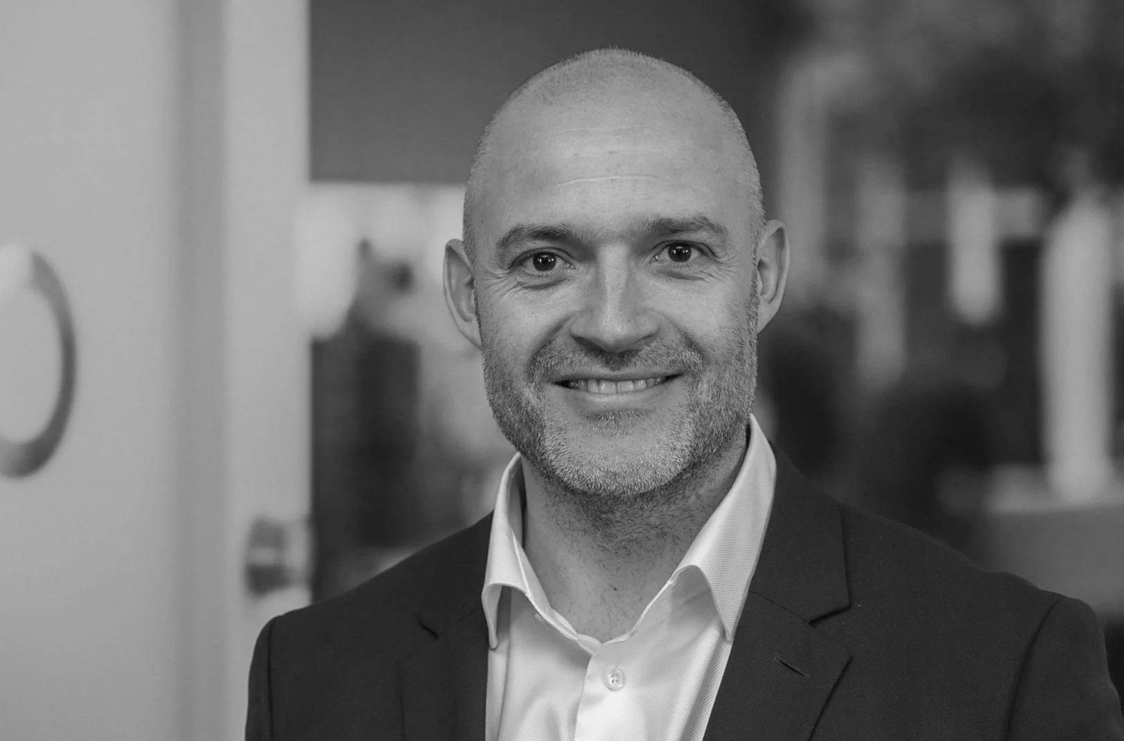 Black and white photo of a smiling man with a shaved head and beard, wearing a suit and white shirt, in an indoor setting.