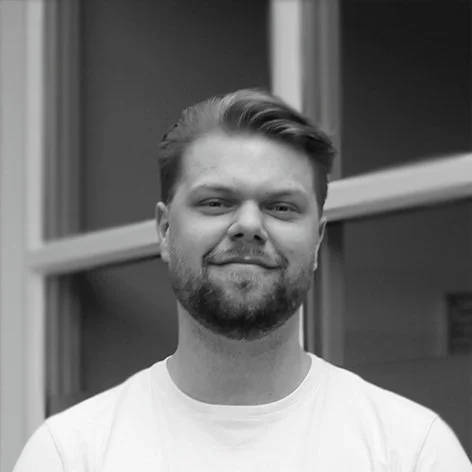 A young man with a beard and short hair, smiling, in front of a window.