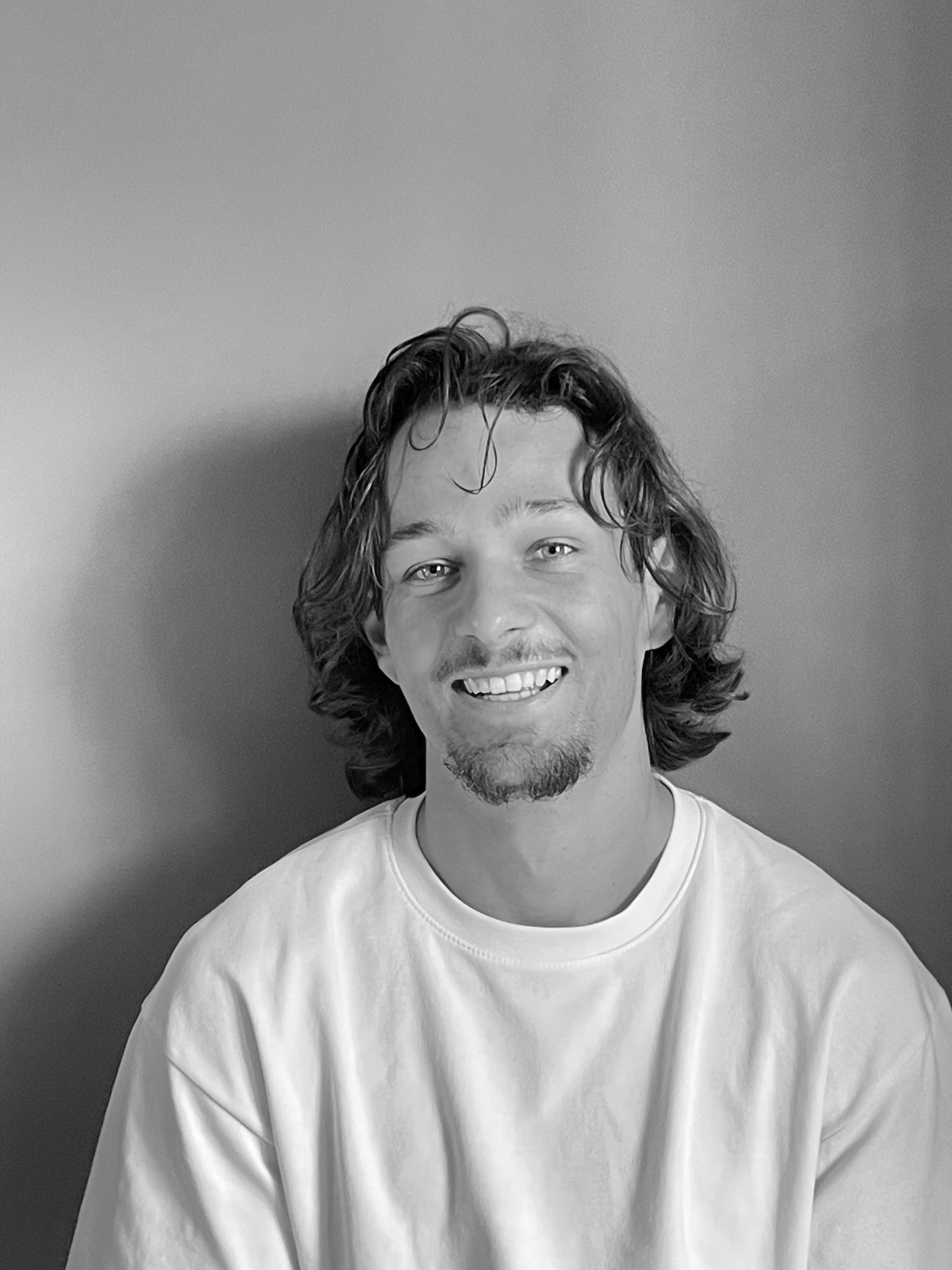 Black and white photograph of a young man with medium-length wavy hair, a light beard, and mustache. He is smiling with visible teeth, wearing a plain white t-shirt, standing against a plain wall background.