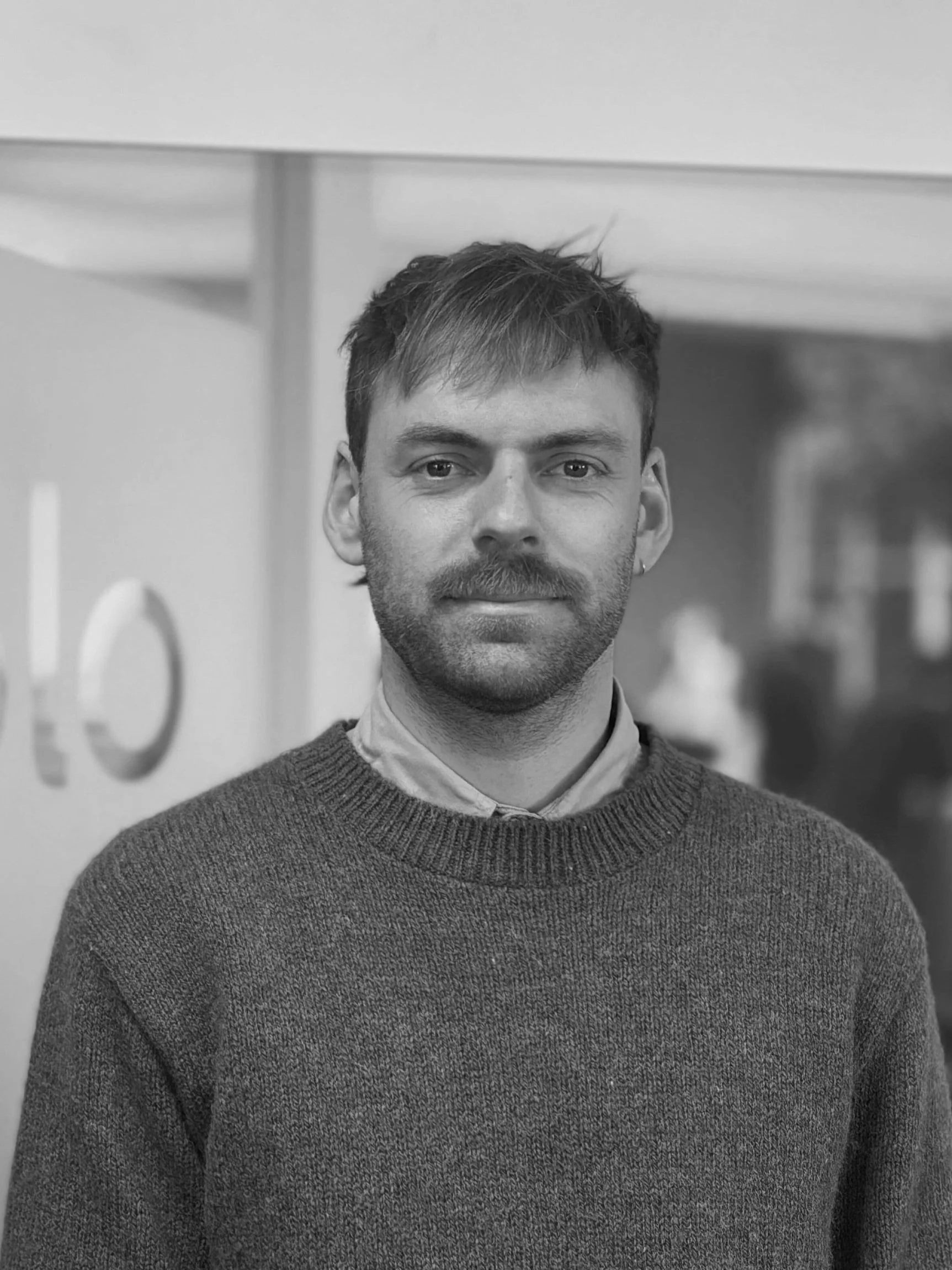 A black-and-white photo of a man with short hair, a beard, and mustache, wearing a sweater over a collared shirt, standing indoors in front of a background with blurred signage.