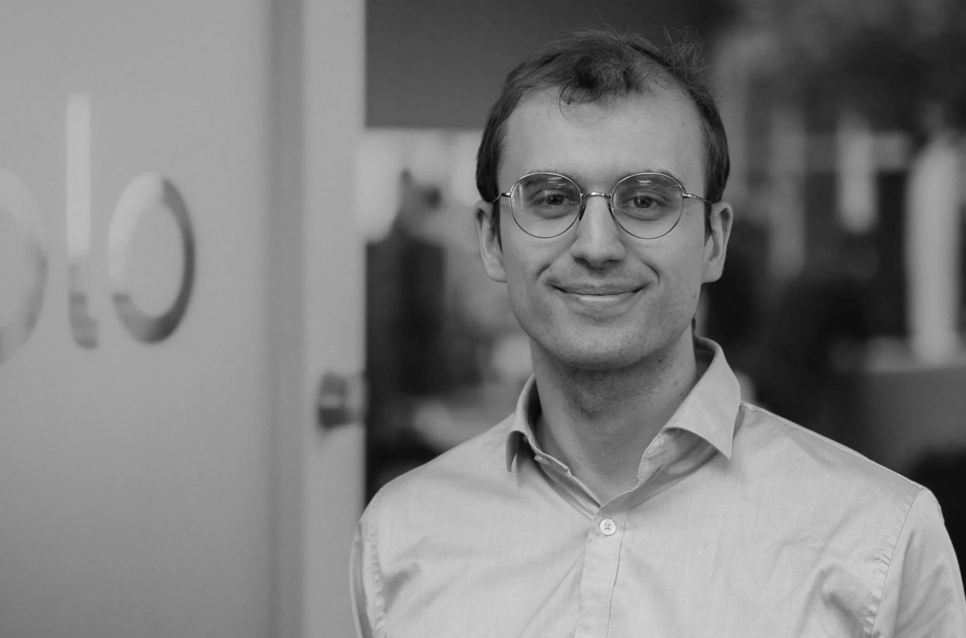 A young man with glasses, a slight smile, wearing a light-colored collared shirt, standing in a hallway with blurred background and a partially visible wall with the number 600.