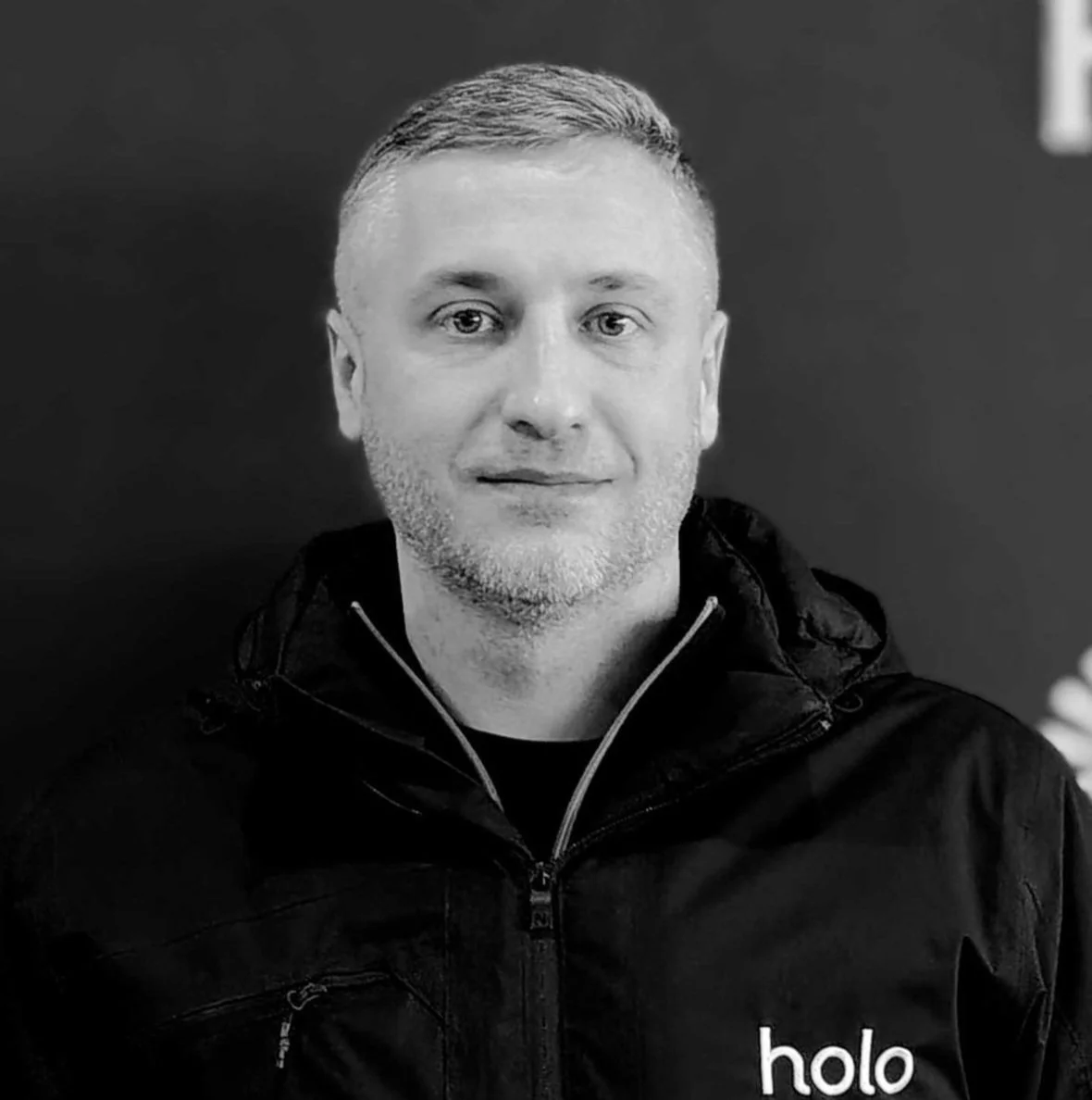 Black and white photo of a man with short hair, light beard, wearing a black jacket with a zipper and the word "holo" on it, standing against a dark background.